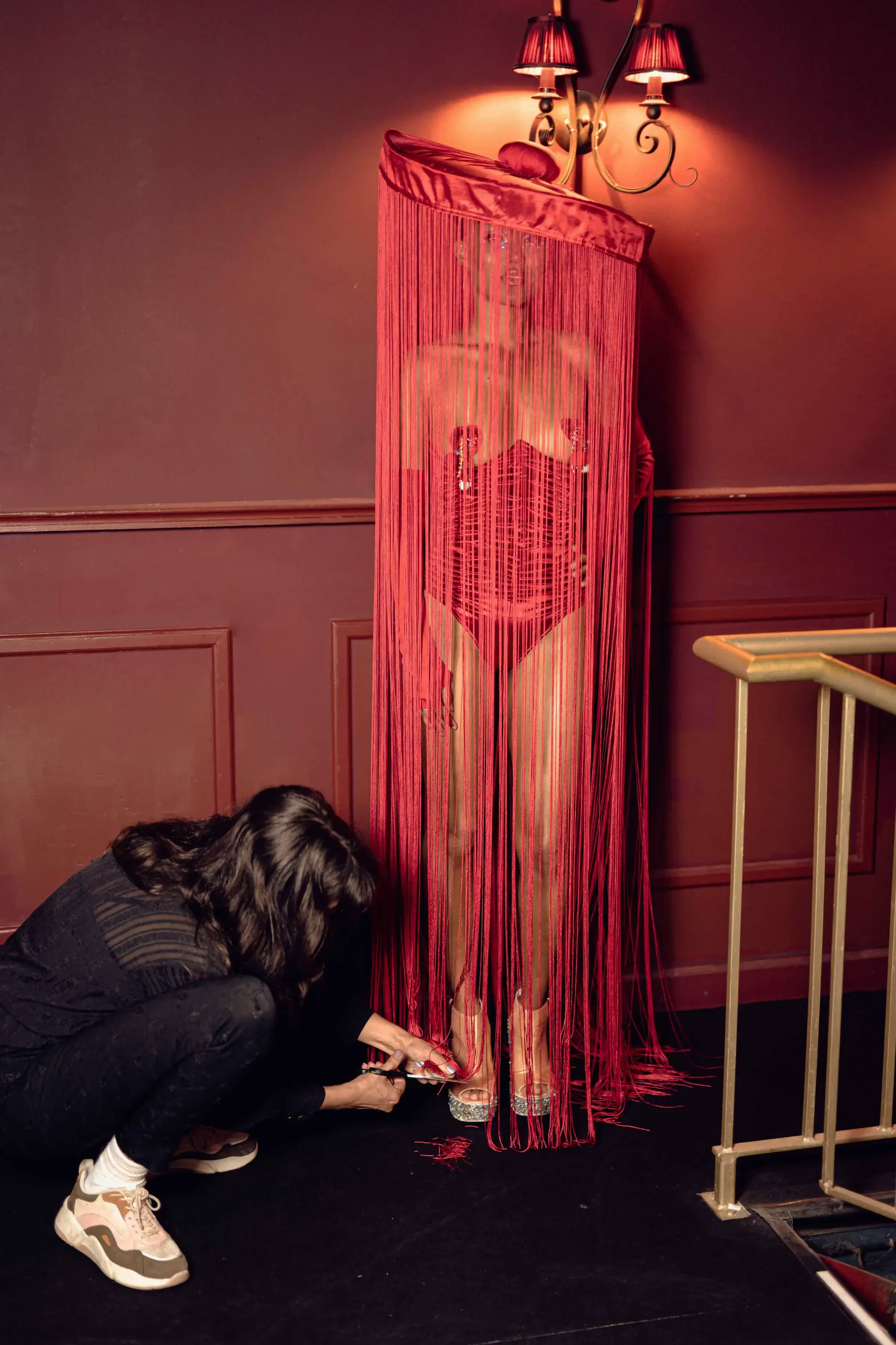 Een vrouw knielt voor een accuraat standbeeld van een vrouw, bedekt met rode draden en een doorzichtige cilinder, in een kamer met borduurwerk op de muur en een muurlamp.