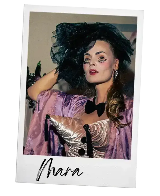 Een vrouw in een extravagante outfit met een grote zwarte muts en make-up, met de naam Mara onderaan de foto.