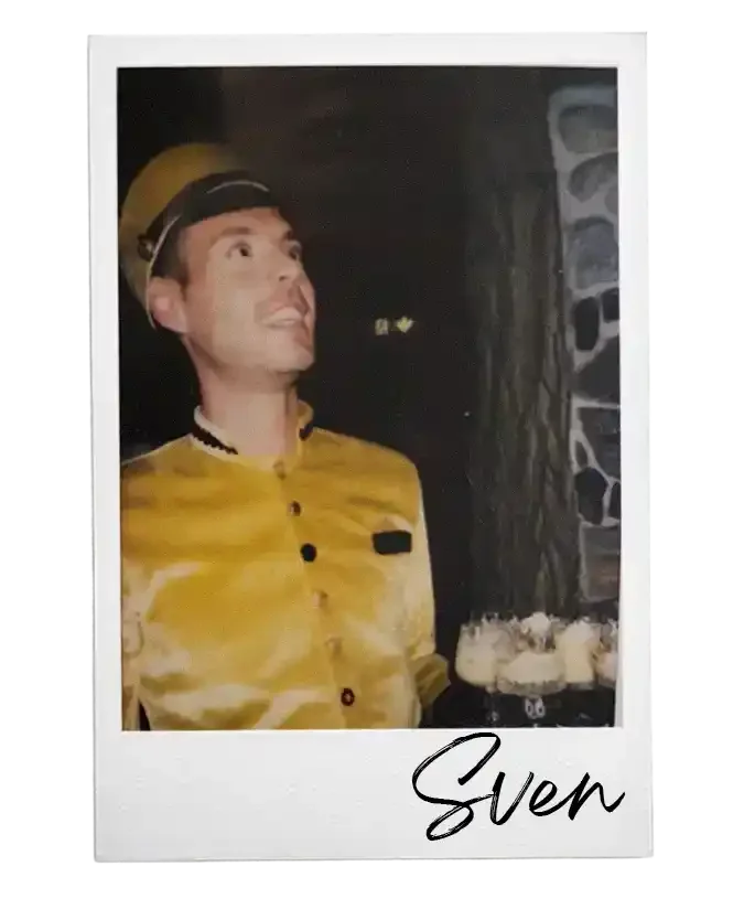 Man in gele uniform met pet en glazen, glimlachend, met een glas met een drankje in de hand, staand voor een stenen muur, ondertekening 'Sven'.