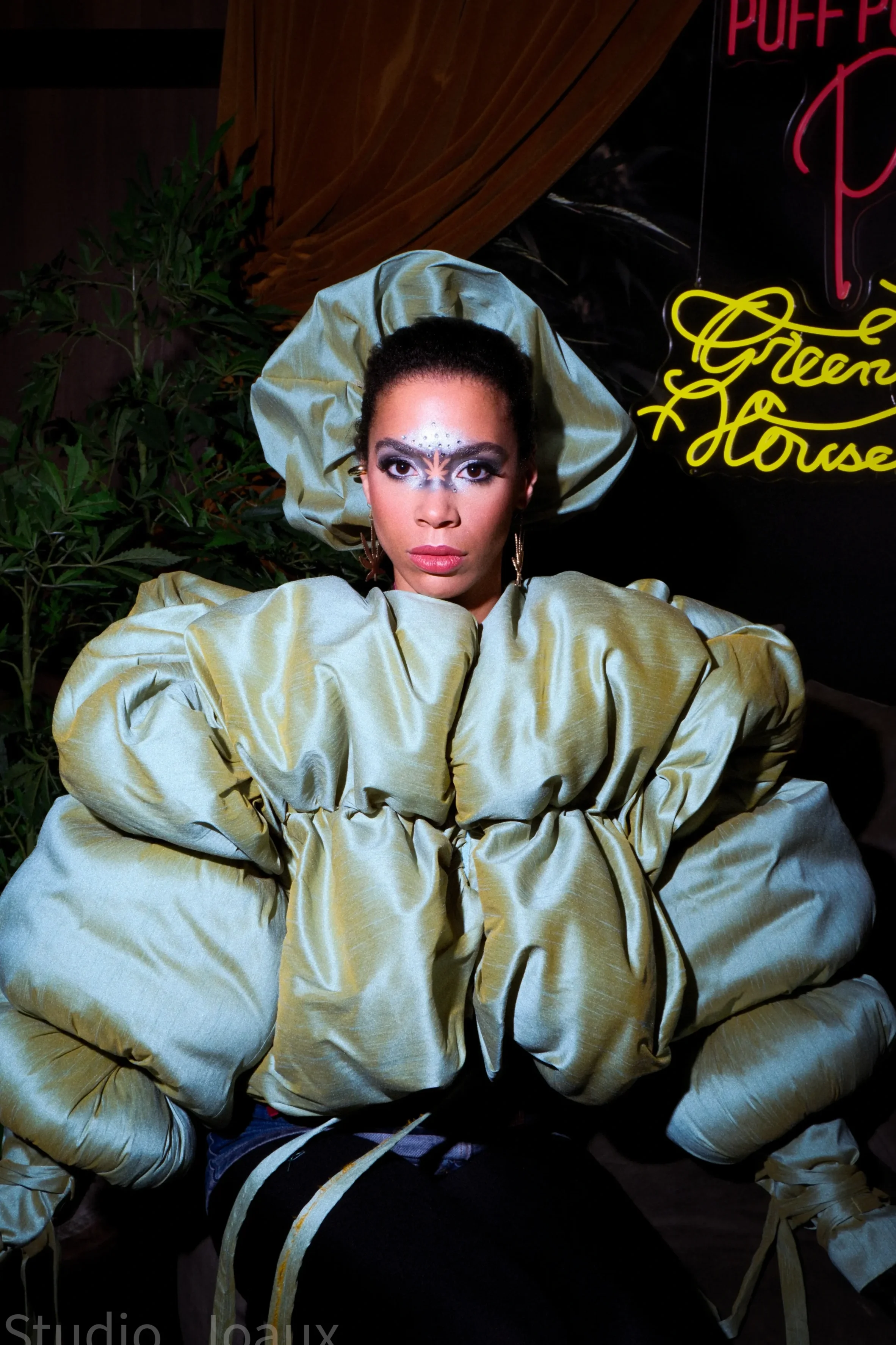 Een vrouw in een opvallende gele jas en grote hoed, met kunstzinnig make-up rond haar ogen, poseert voor een achtergrond met planten en neonlichten die onder andere de woorden 'Puff Puff' en 'Green & Horse' bevatten.