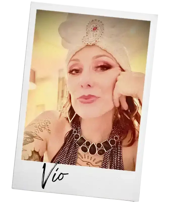 Een vrouw met make-up en een hoofddoek, die op een foto posert met haar hoofd ondersteund door haar hand. Ze draagt grote oorbellen en een sierlijke ketting. De naam 'Vio' is onderaan de foto geschreven.