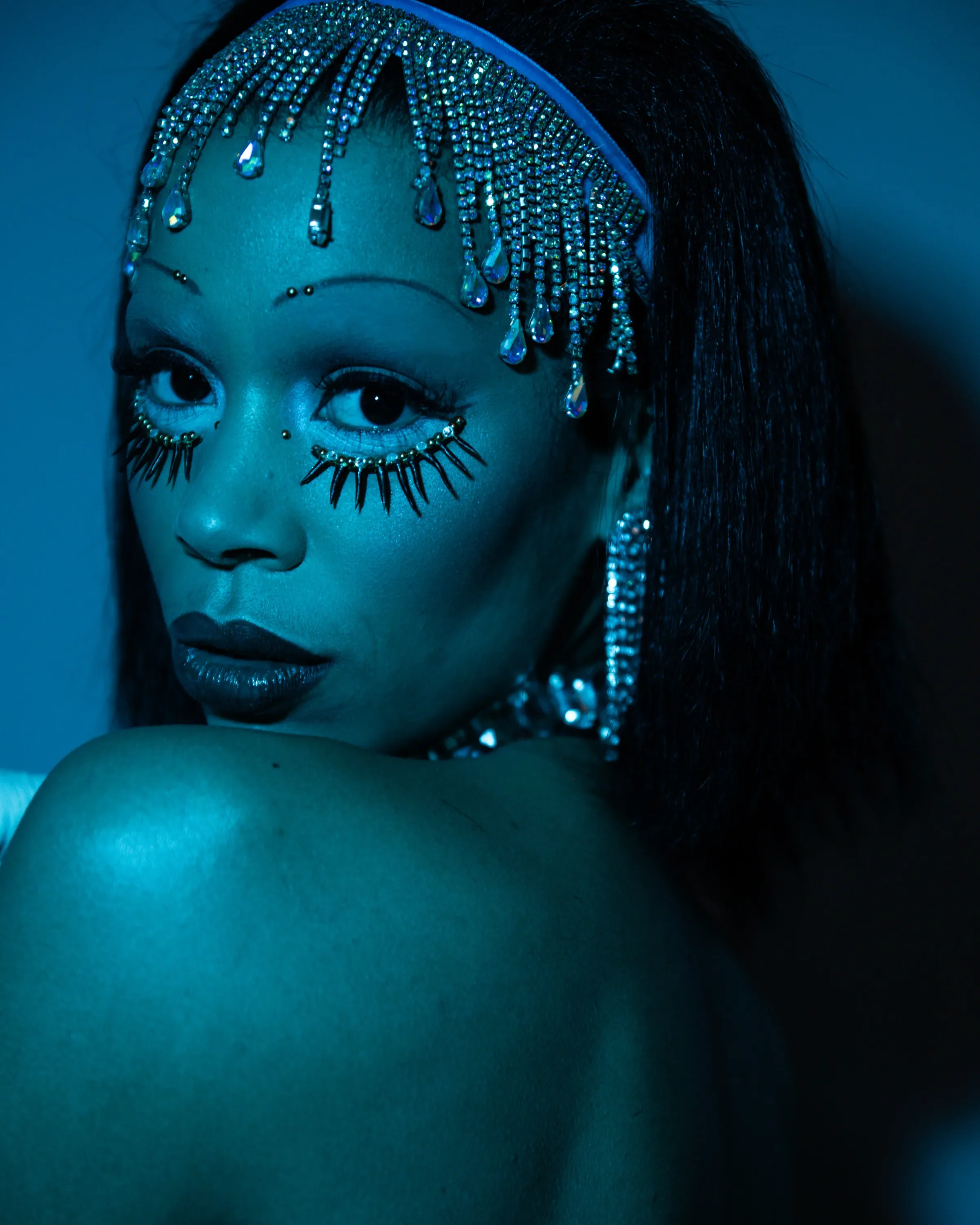 Portret van een vrouw met opvallende make-up en sieraden, op een blauwe achtergrond.