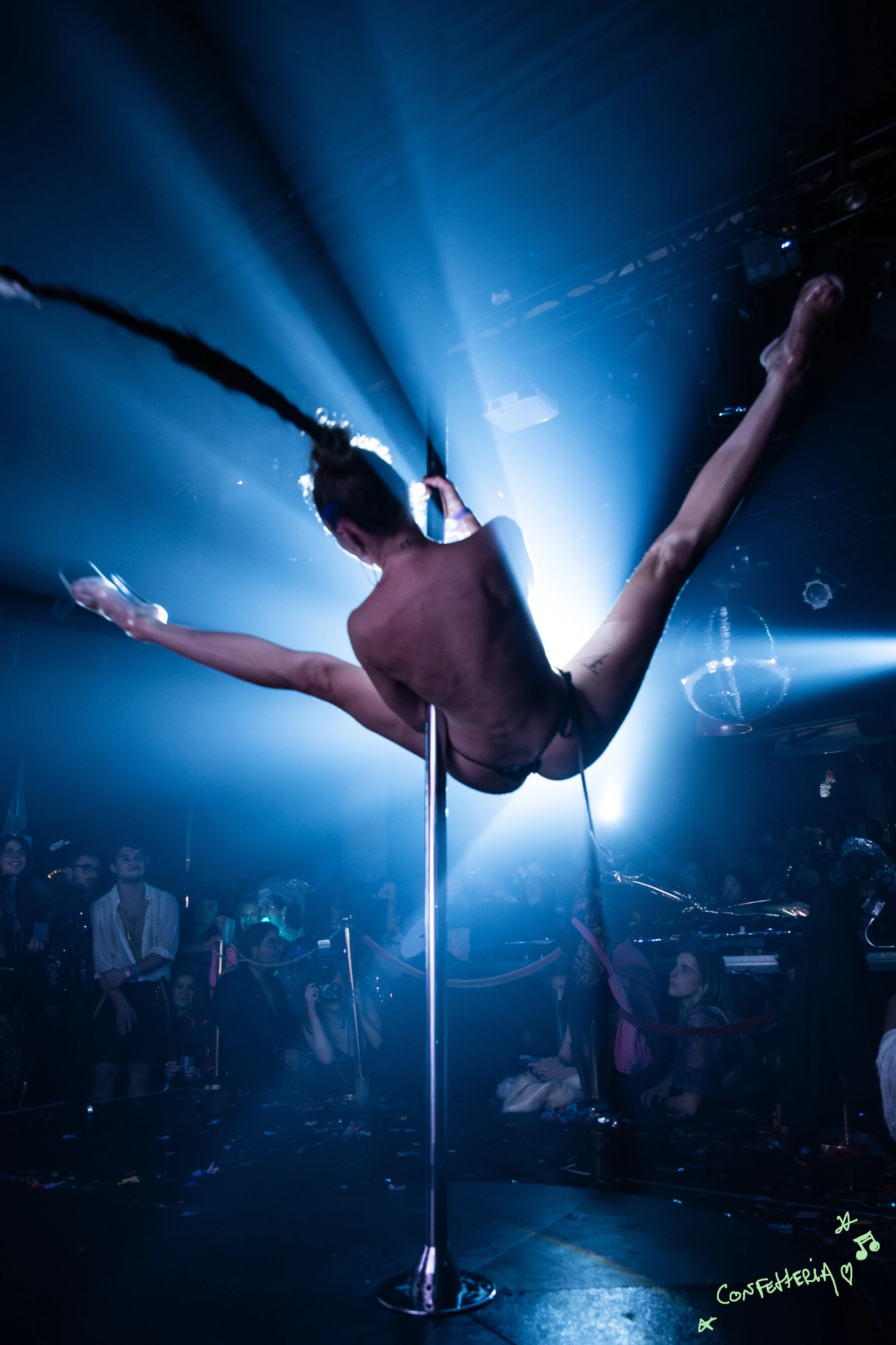 Een danseres voert een acrobatische pole dance uit op een podium in een club, met een publiek dat toekijkt. Er zijn blauwe lichten en discoballen op de achtergrond.
