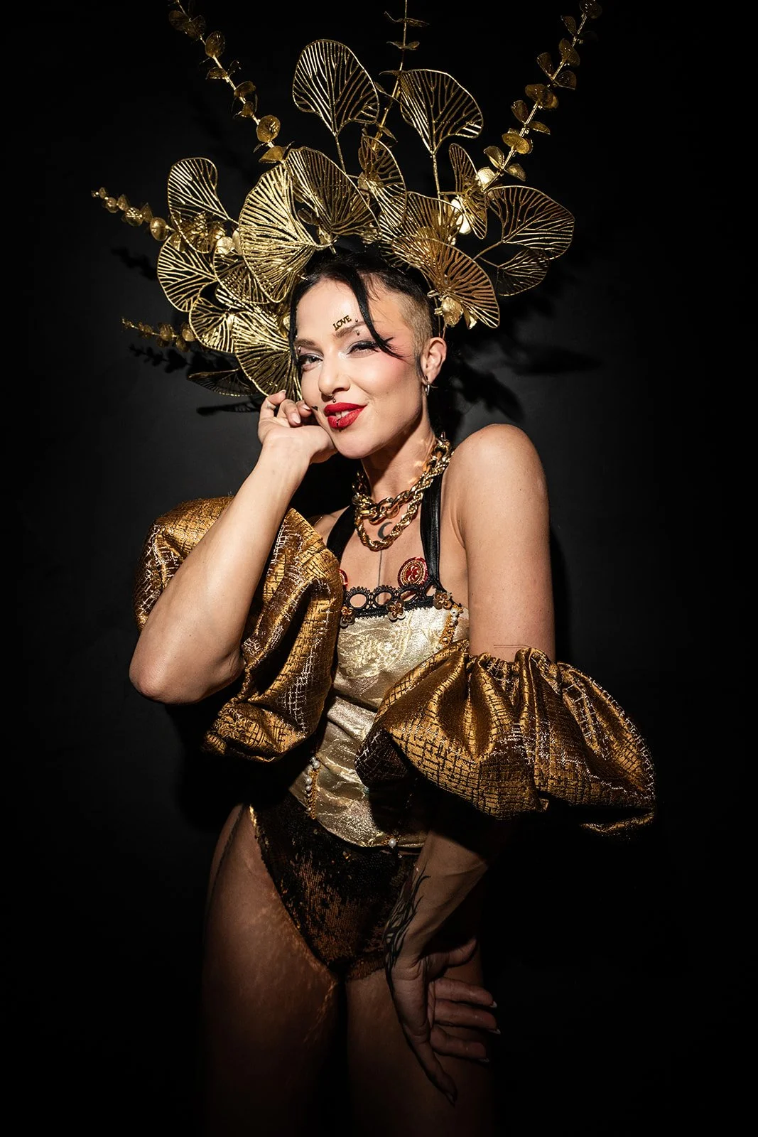 Vrouw in een glanzend goudkleurig kledingstuk met een grote gouden hoed en sieraden, poseert tegen een zwarte achtergrond.