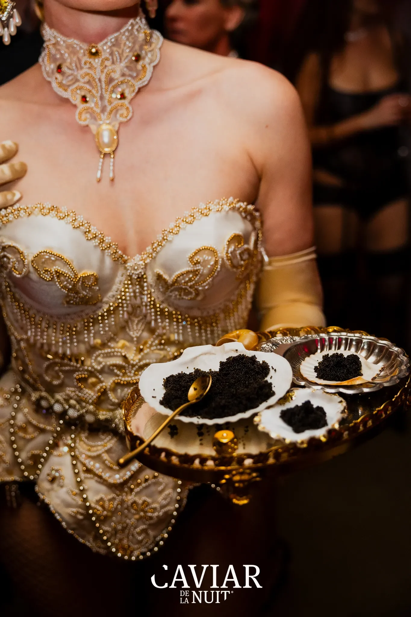 Vrouw in een sierlijk, gouden en parelversierd jurk met een kanten ketting die parels en een groot parel in het midden draagt, houdt een zilveren schaal met zwarte kaviaar in een luxe setting.