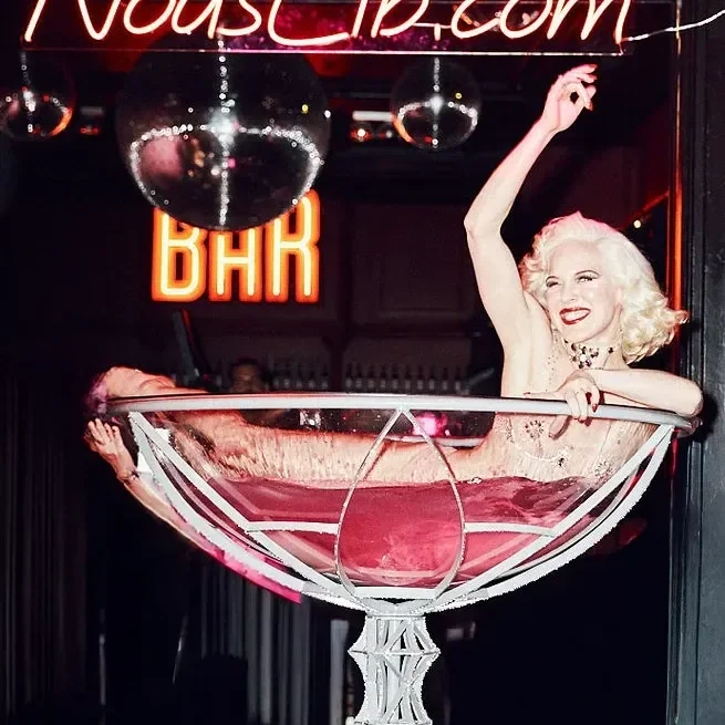 Vrouw met blonde haar in een grote cocktailglas met roze drank, dansend en glimlachend in een bar met neonverlichting.