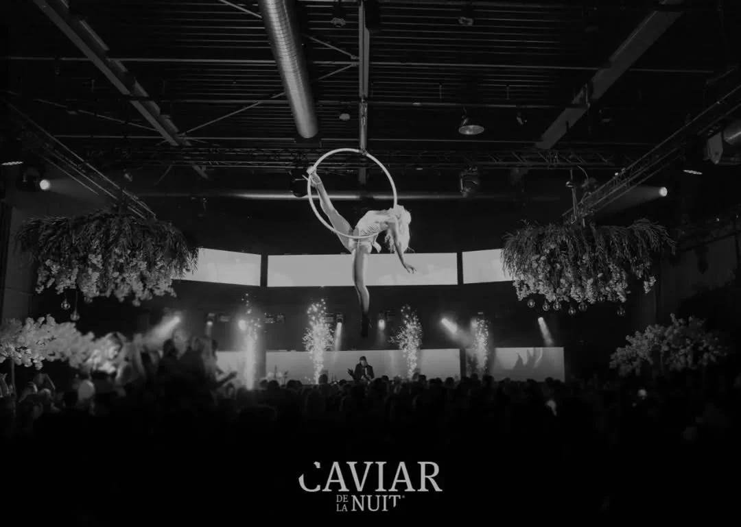 Een acrobaat beweegt zich aan een cirkel ronddraaiend boven een publiek in een concertzaal met bloemendecoraties.