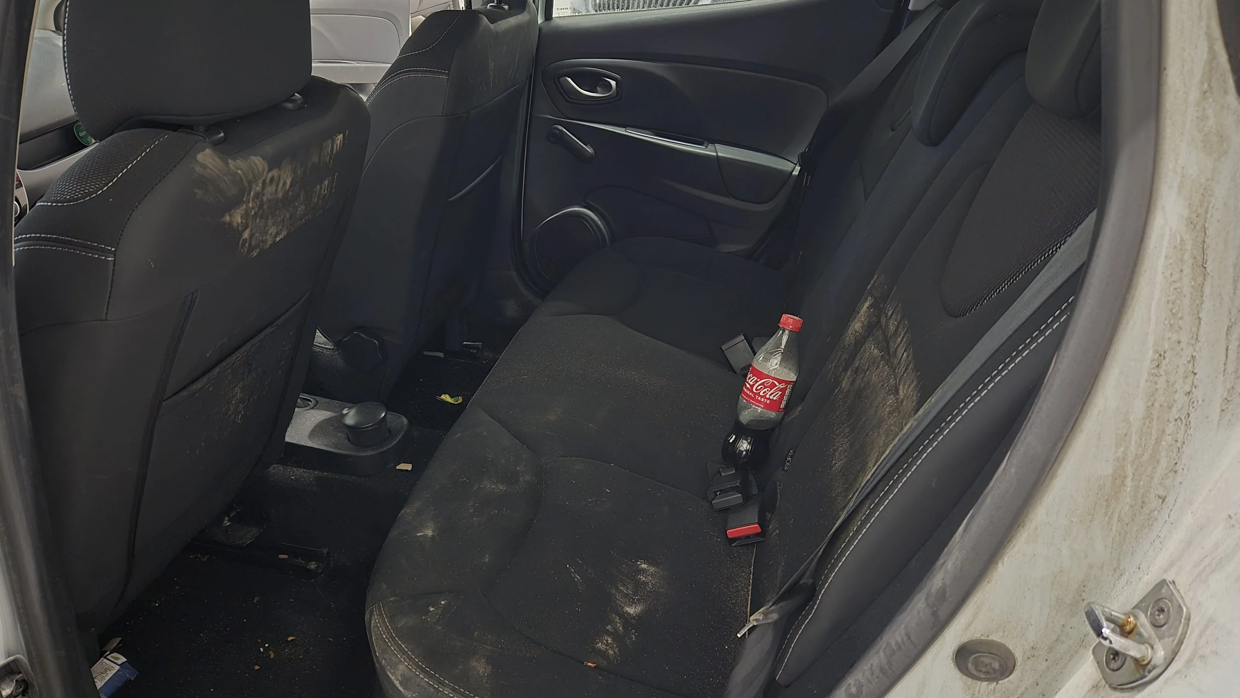 Bild av baksätet i en bil med smuts på golvet och ett tomt Coca-Cola-flaska i sätet.