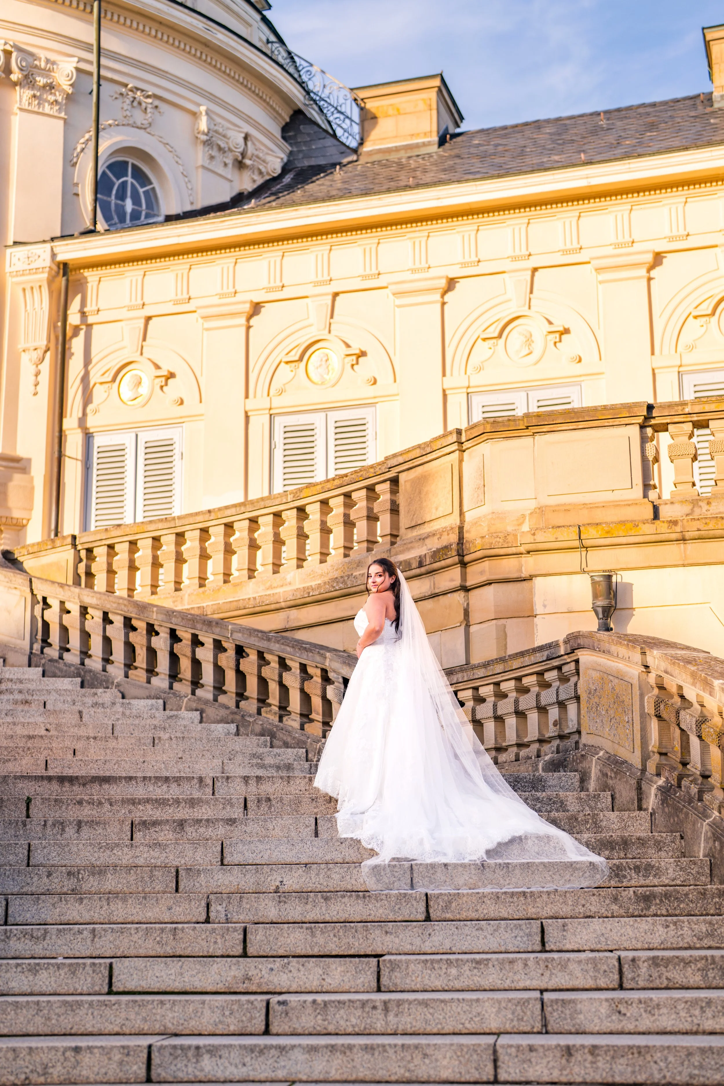 Braut in Hochzeitskleid auf Treppen vor einem historischen Gebäude bei Sonnenuntergang.