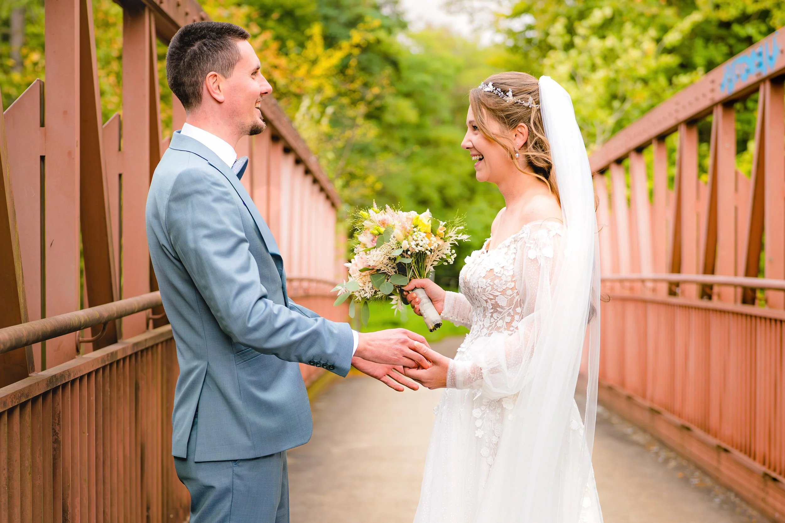 Ein Brautpaar beim Standesamt, das sich die Hände reicht und sich gegenseitig anlacht, im Hintergrund grüne Bäume.
