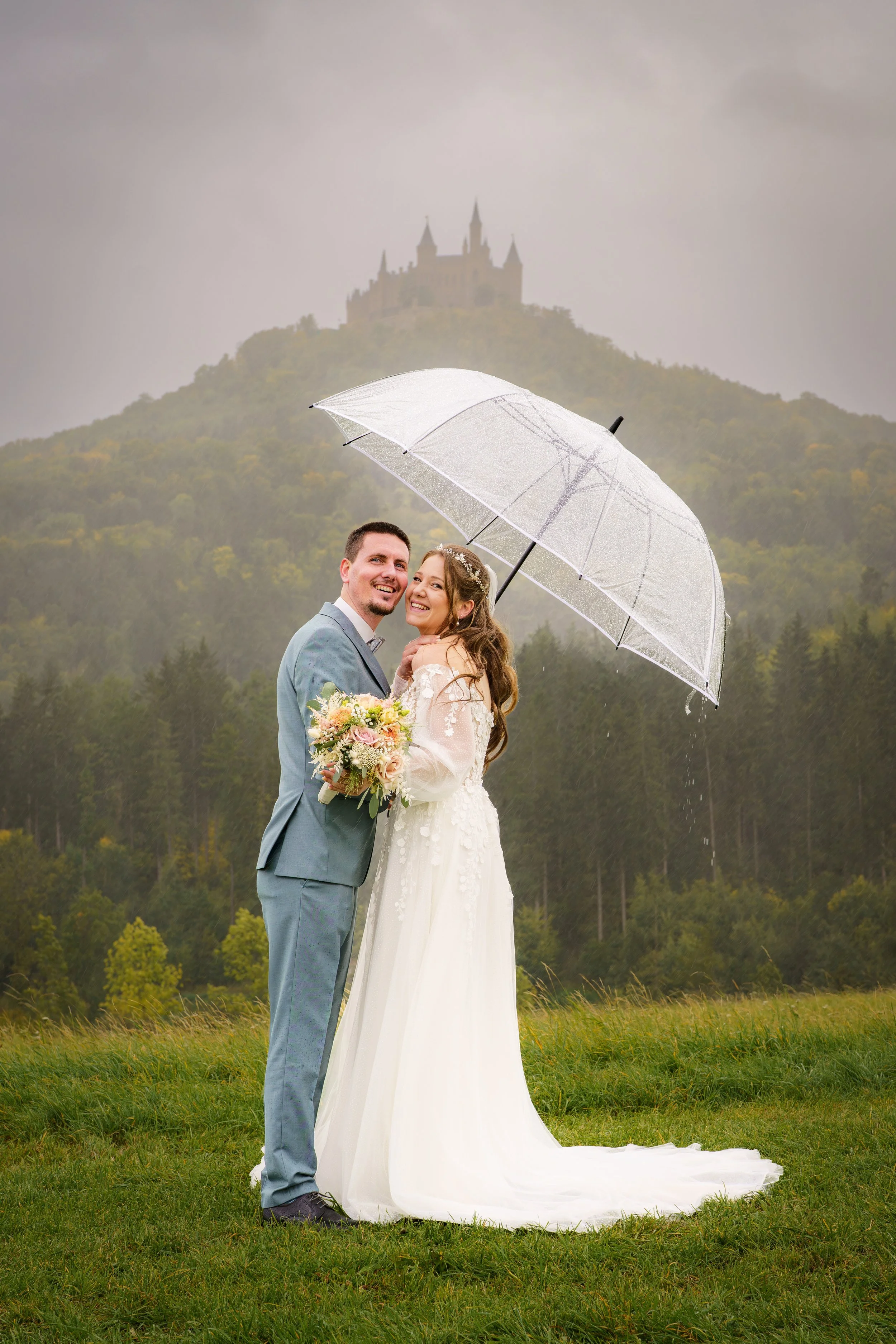 Ein Hochzeitsfoto eines Paares, das bei Regen draußen steht, das Brautpaar unter einem transparenten Regenschirm, im Hintergrund ein Schloss auf einem Hügel, umgeben von Wald, auf einer Wiese.