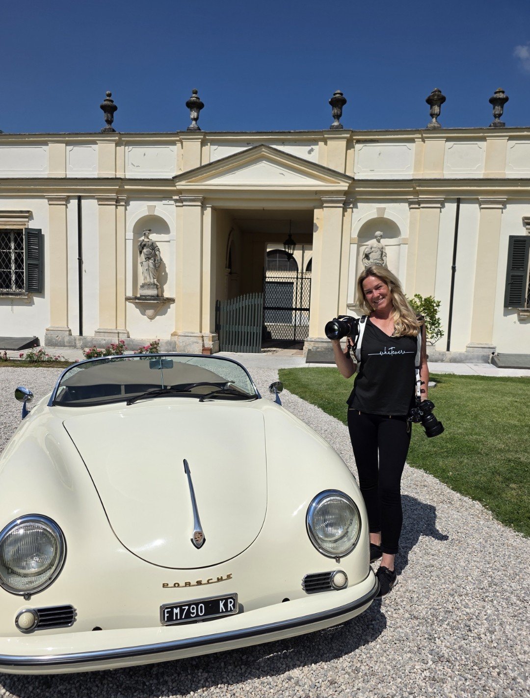 Eine Frau steht neben einem hellgelben Porsche 356, hält zwei Kameras in den Händen und lächelt, im Hintergrund ein historisches Gebäude mit Statuen.