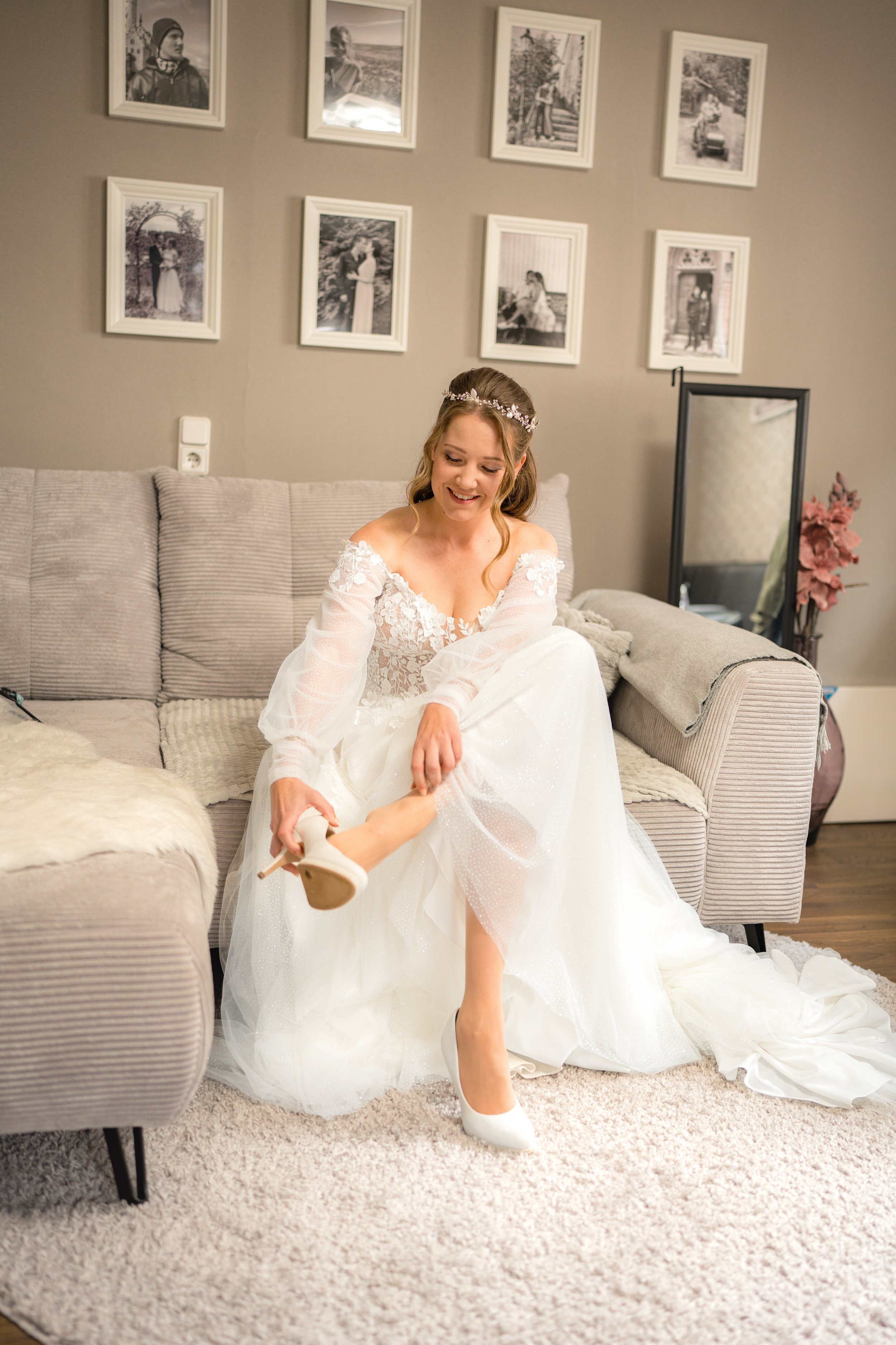 Braut sitzt auf einem Sofa, zieht einen weißen Hochzeits-High Heel Schuh an, lächelt, im Hintergrund eine Wand mit schwarz-weiß Fotos.