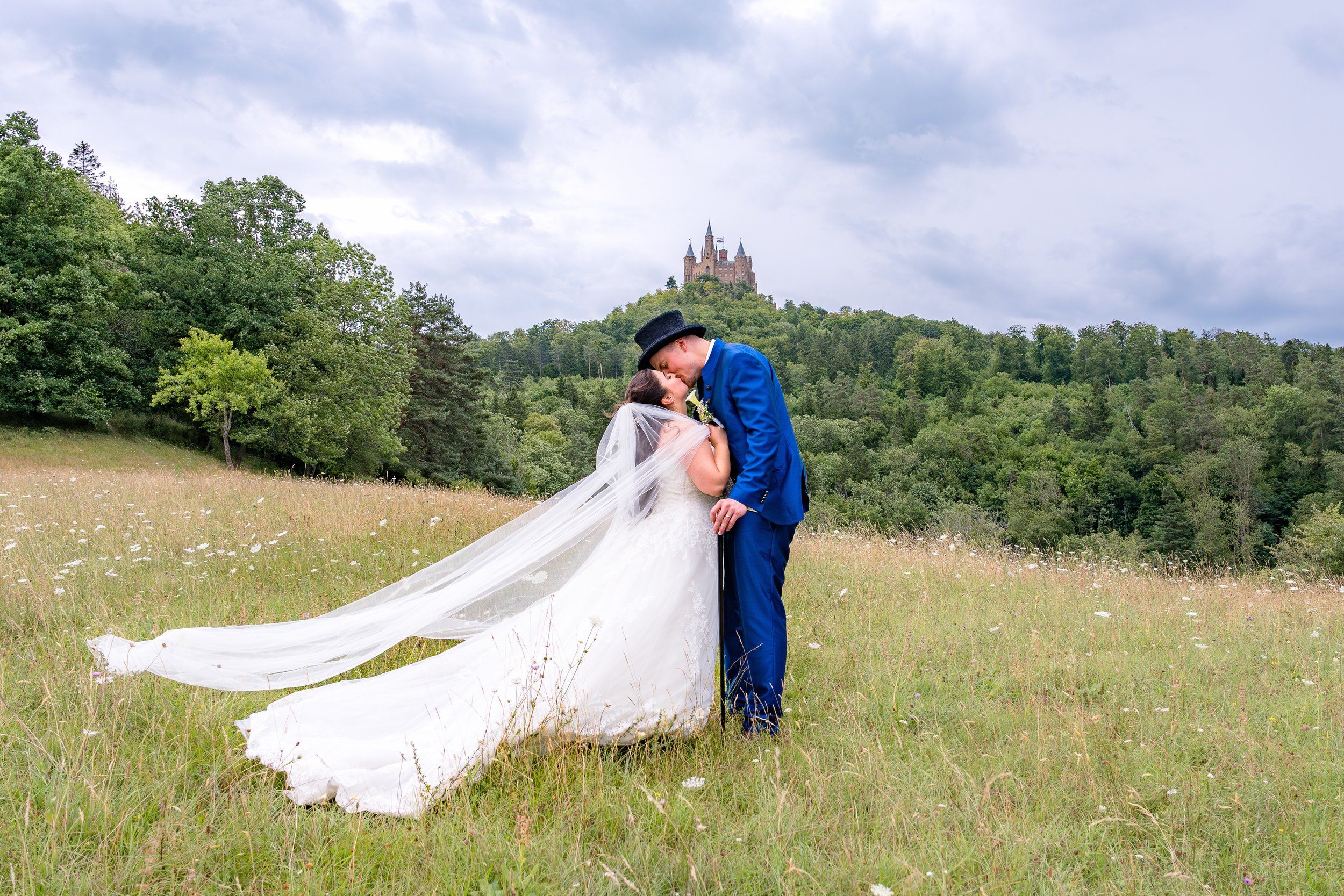 Ein Brautpaar küsst sich unterhalb der Burg Hohenzollern. Sie stehen auf einer Wiese im Hintergrund ist die Burg zu sehen. Sie trägt ein Brautkleid mit Schleier und er einen blauen Anzug.
