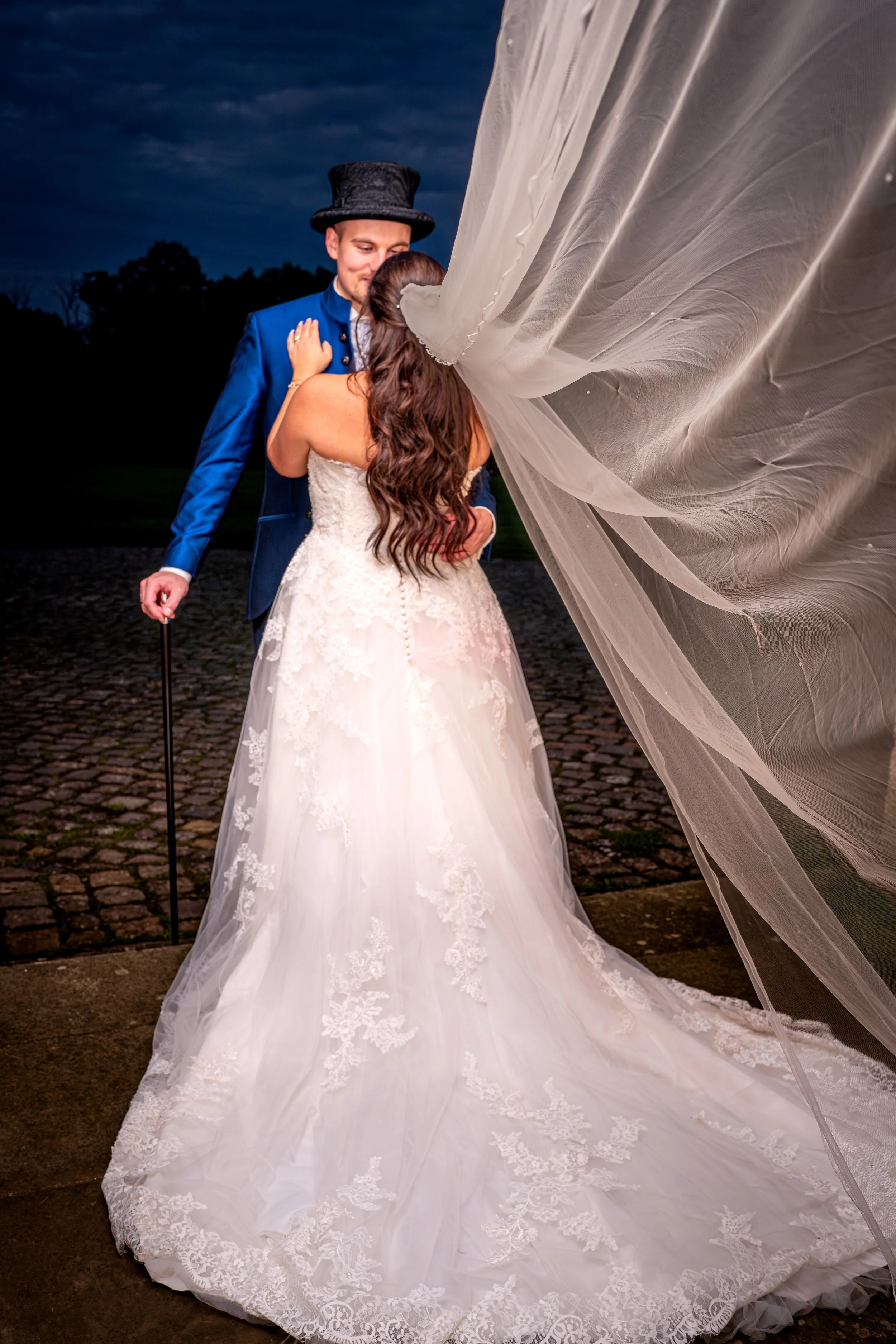 Ein Brautpaar bei Nacht, die Braut trägt ein weißes Spitzenkleid, der Bräutigam trägt einen blauen Anzug mit Zylinder und Gehstock, sie küsst sich unter einem großen Schleier.