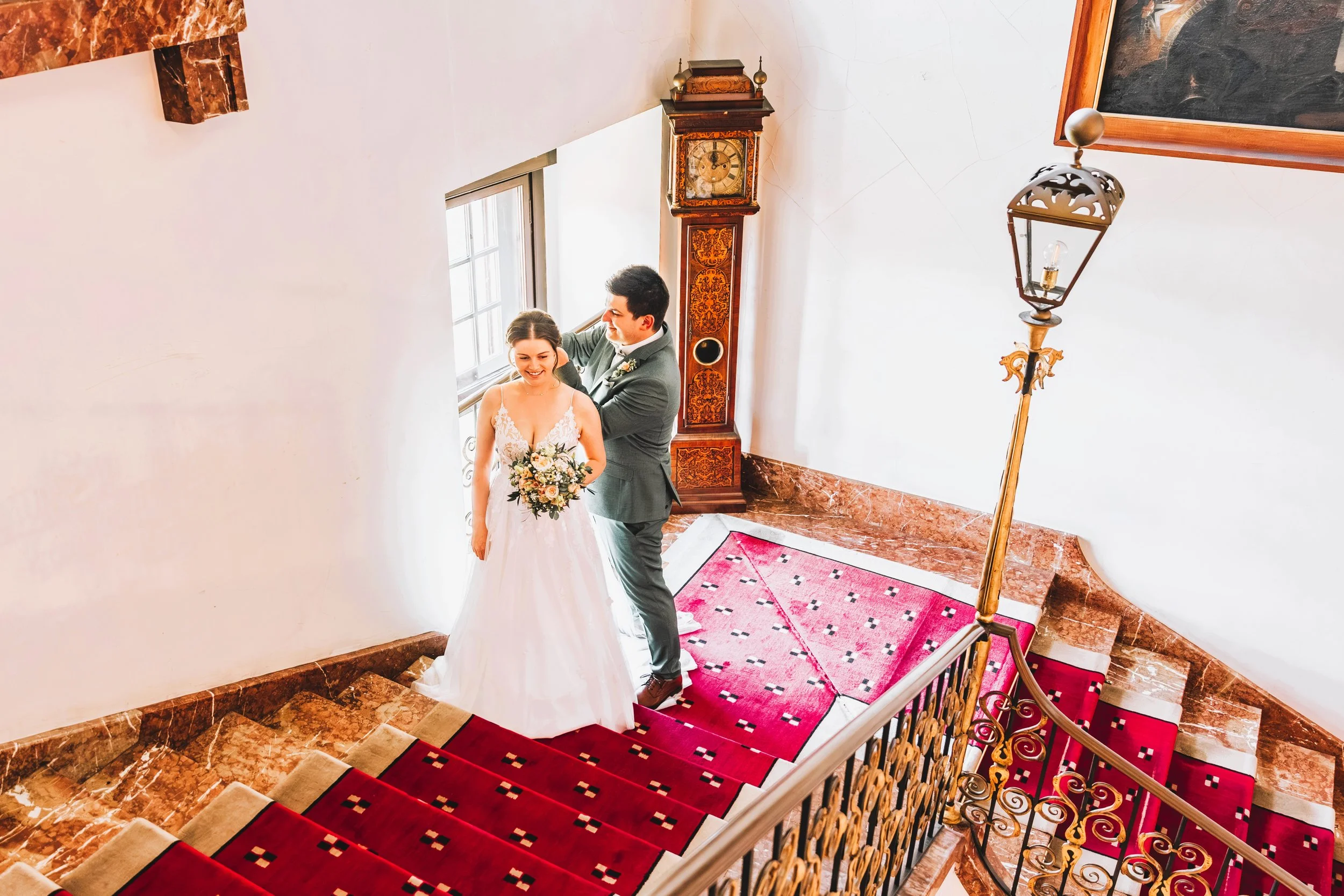 Verliebtes Brautpaar steht auf einer roten Teppichtreppe im Haus, die Braut hält einen Blumenstrauß, das Paar lächelt. Im Hintergrund ein antiker Kamin, eine Standlampe und Bilder an der Wand.