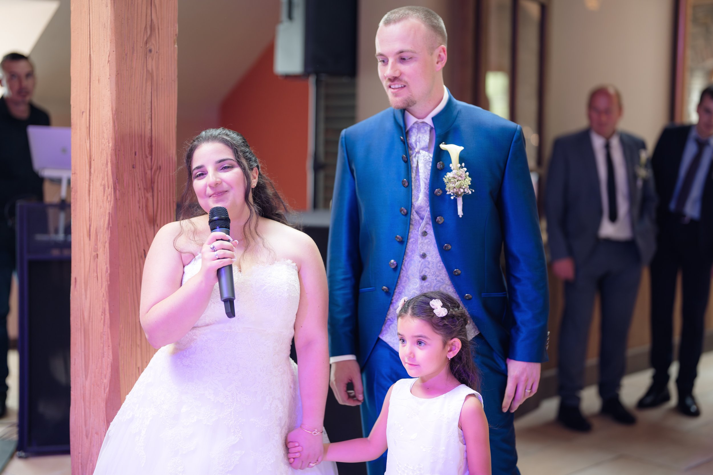 Braut spricht in ein Mikrofon bei ihrer Hochzeit, während der Bräutigam und ein kleines Mädchen Hände halten und zuschauen, im Hintergrund Gäste im Anzug.