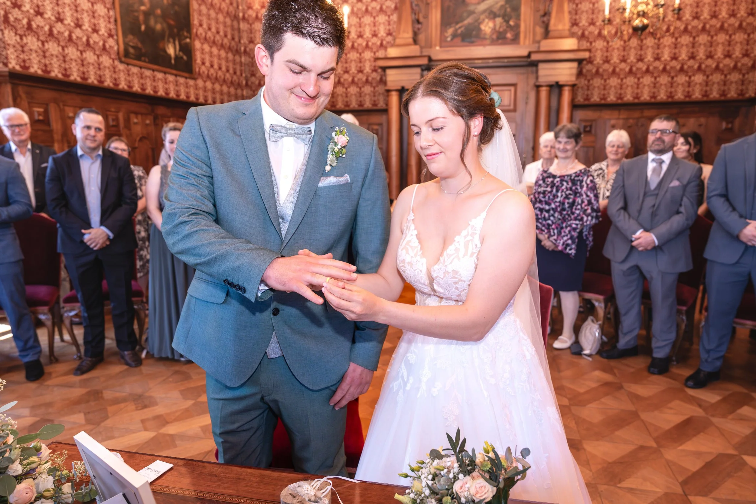 Das Brautpaar tauscht während ihrer Hochzeit Ringe aus, umgeben von Gästen in einem festlichen Raum mit Holzverkleidung und Gemälden an den Wänden.