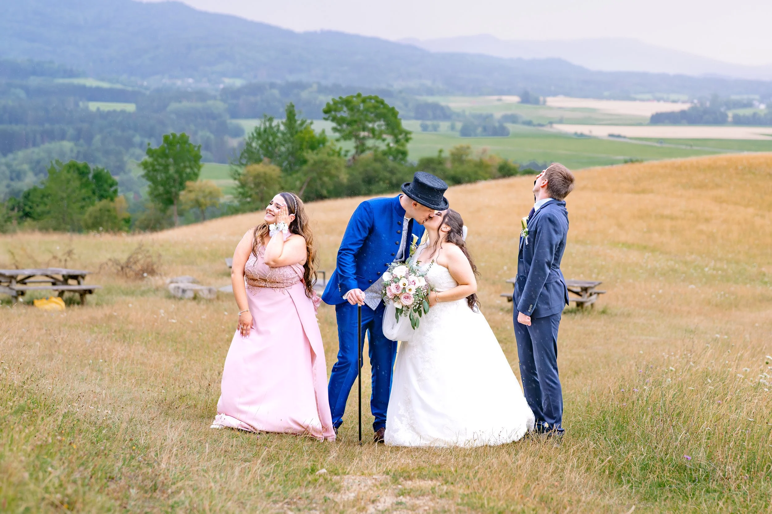 Hochzeit im Freien mit vier Personen, einem Brautpaar, einem Mann im blauen Anzug und einem weiteren Mann im dunklen Anzug, bei einer Umarmung, in einer ländlichen Landschaft mit Hügeln und Bäumen im Hintergrund.