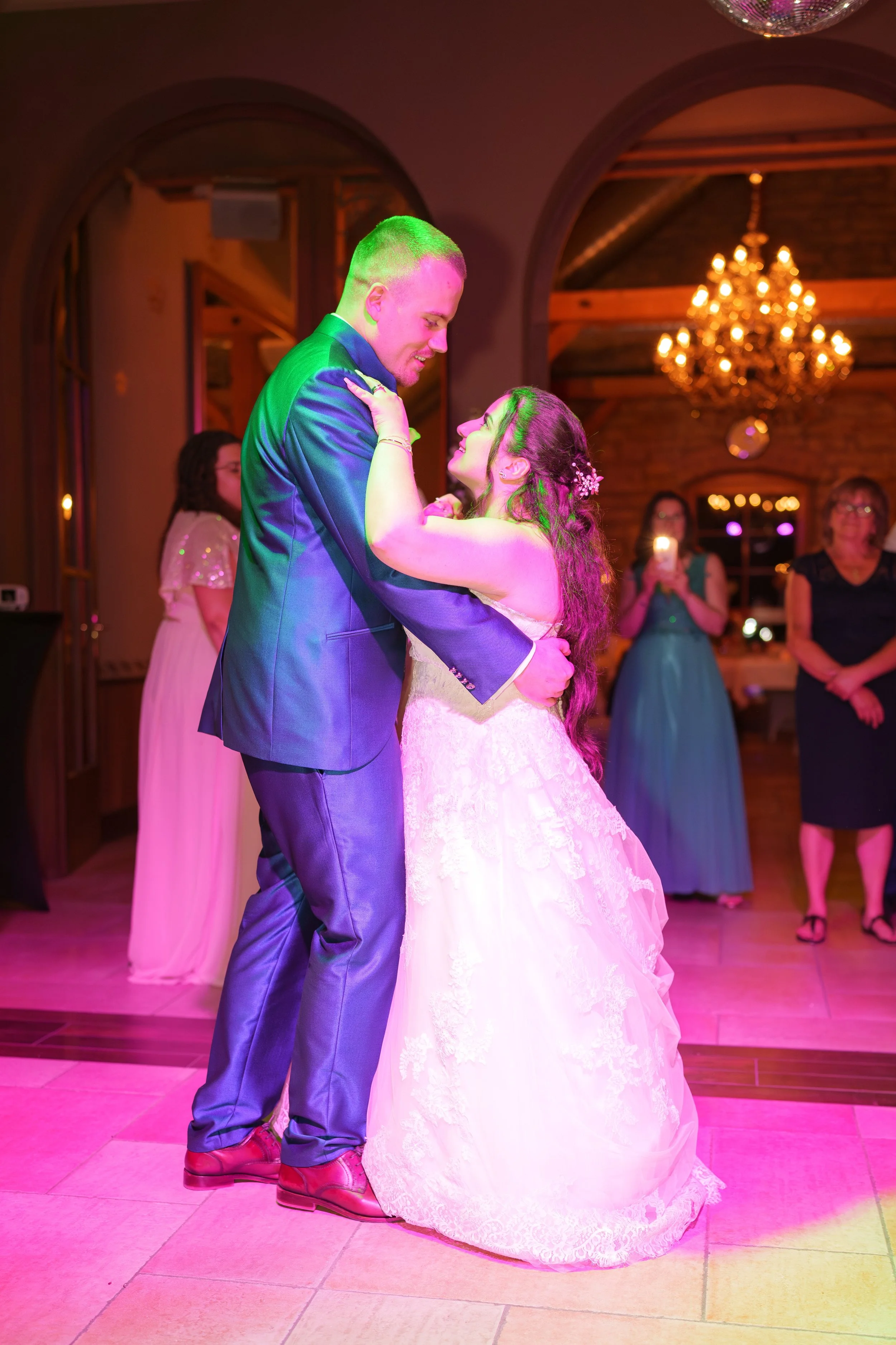 Ein Brautpaar tanzt auf einer Hochzeit, der Bräutigam trägt einen dunklen Anzug, die Braut ein weißes Brautkleid, im Hintergrund feiern Gäste und eine goldene Kronleuchter