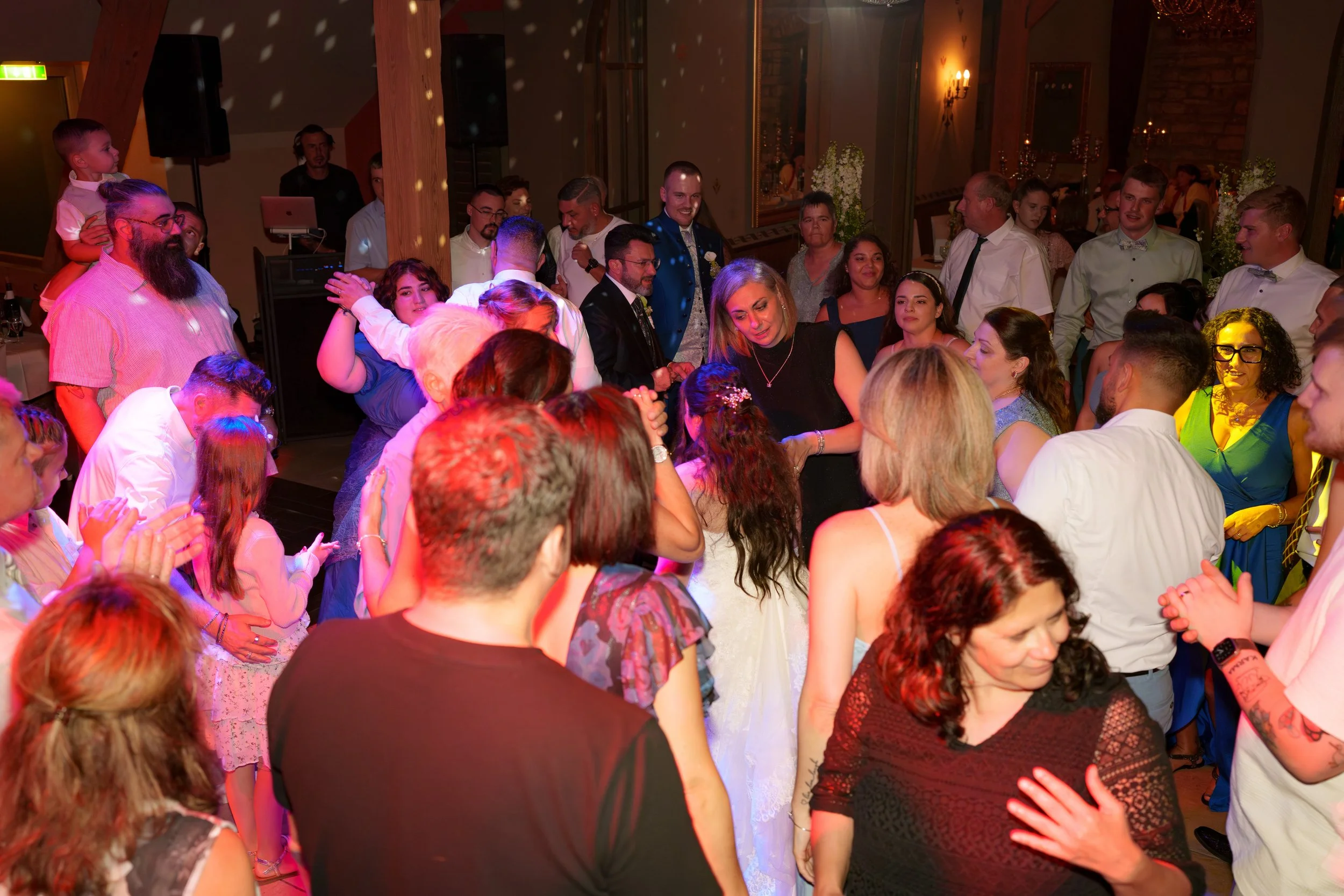 Menschen tanzen und feiern in einem festlich dekorierten Raum mit warmem Licht und Blumen, vermutlich bei einer Hochzeit.