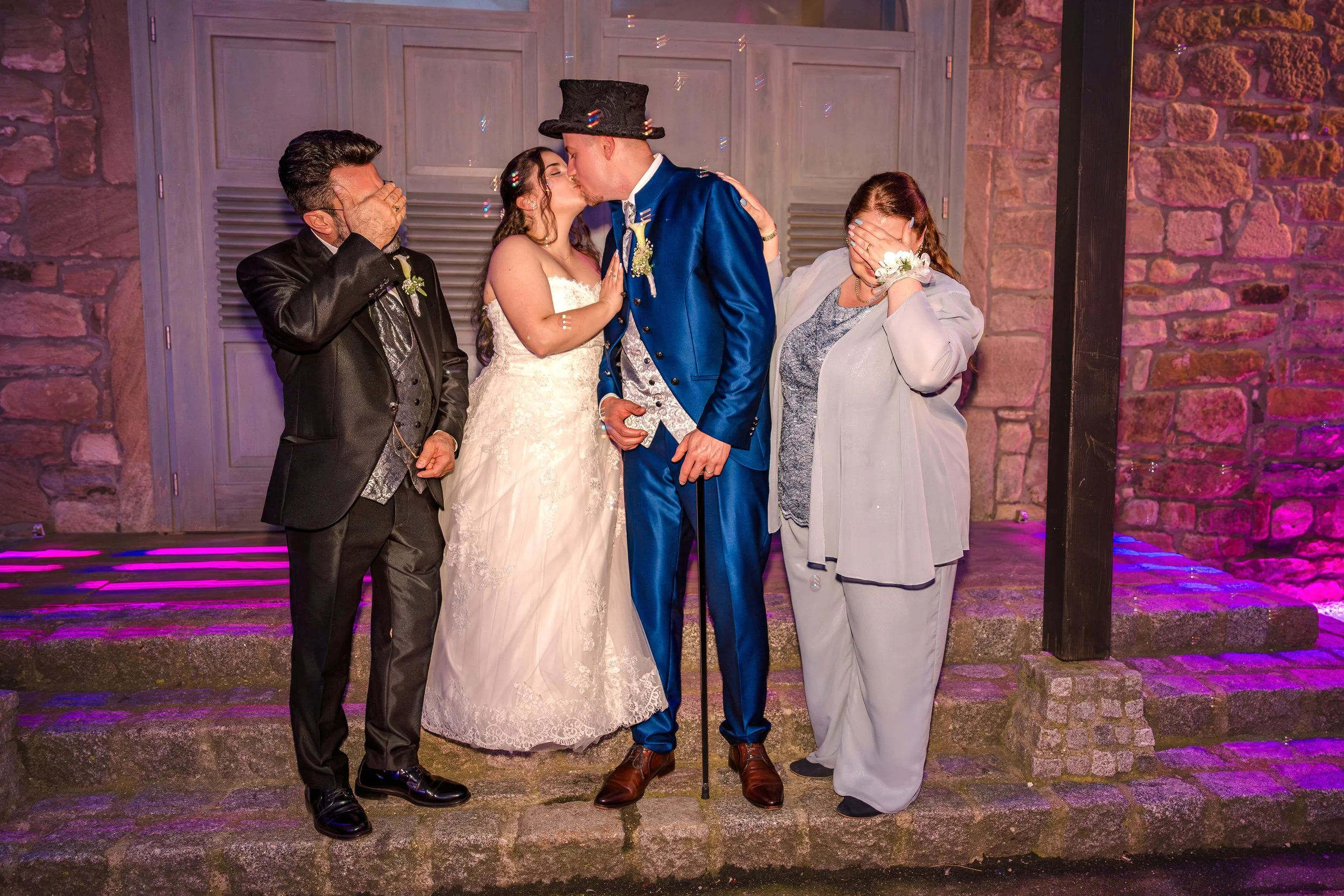 Jubelnde Hochzeitspaar beim Küssen, flankiert von zwei lachenden Frauen, vor einer Steinwand mit bunten Lichtern.