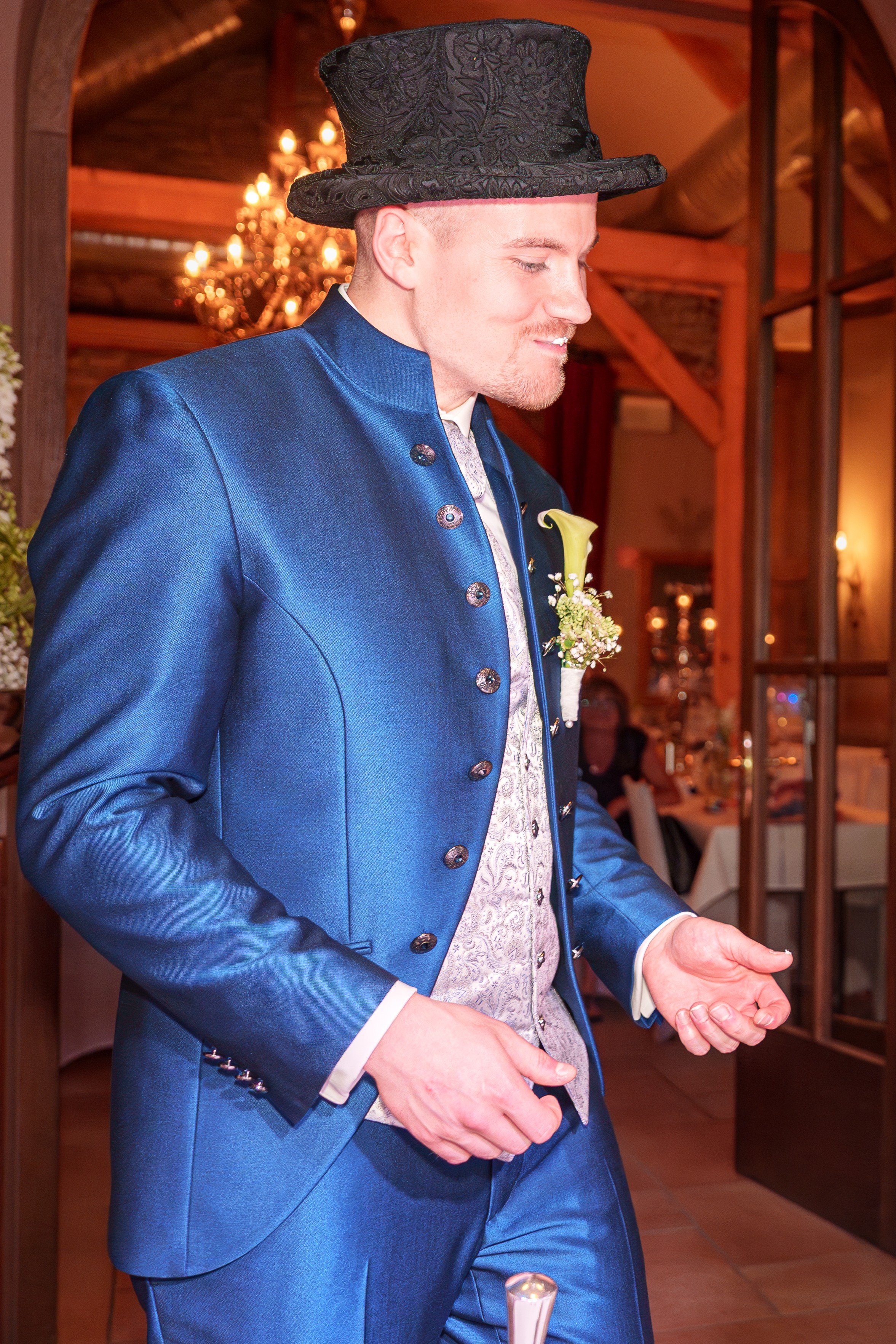 Ein Mann in einer eleganten blauen Jacke und einer erhaltenen Zylinderhut beim Auftritt auf einer Hochzeit oder Feier, im Hintergrund ein festlicher Raum mit Kerzen und Dekoration.