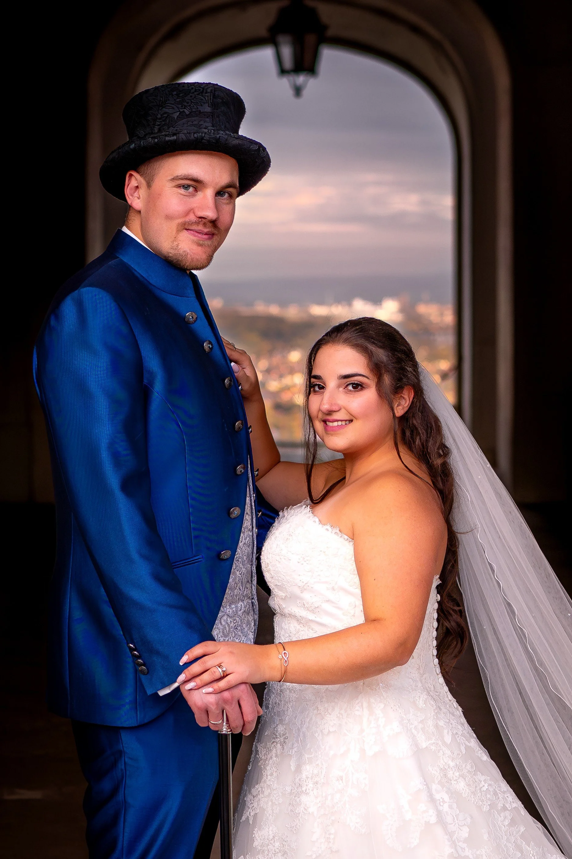 Ein Brautpaar posiert an einem Fenster mit Blick auf die Stadt im Hintergrund, die Braut in einem weißen Hochzeitskleid mit Schleier, der Bräutigam in einem blauen Smoking mit Zylinder.