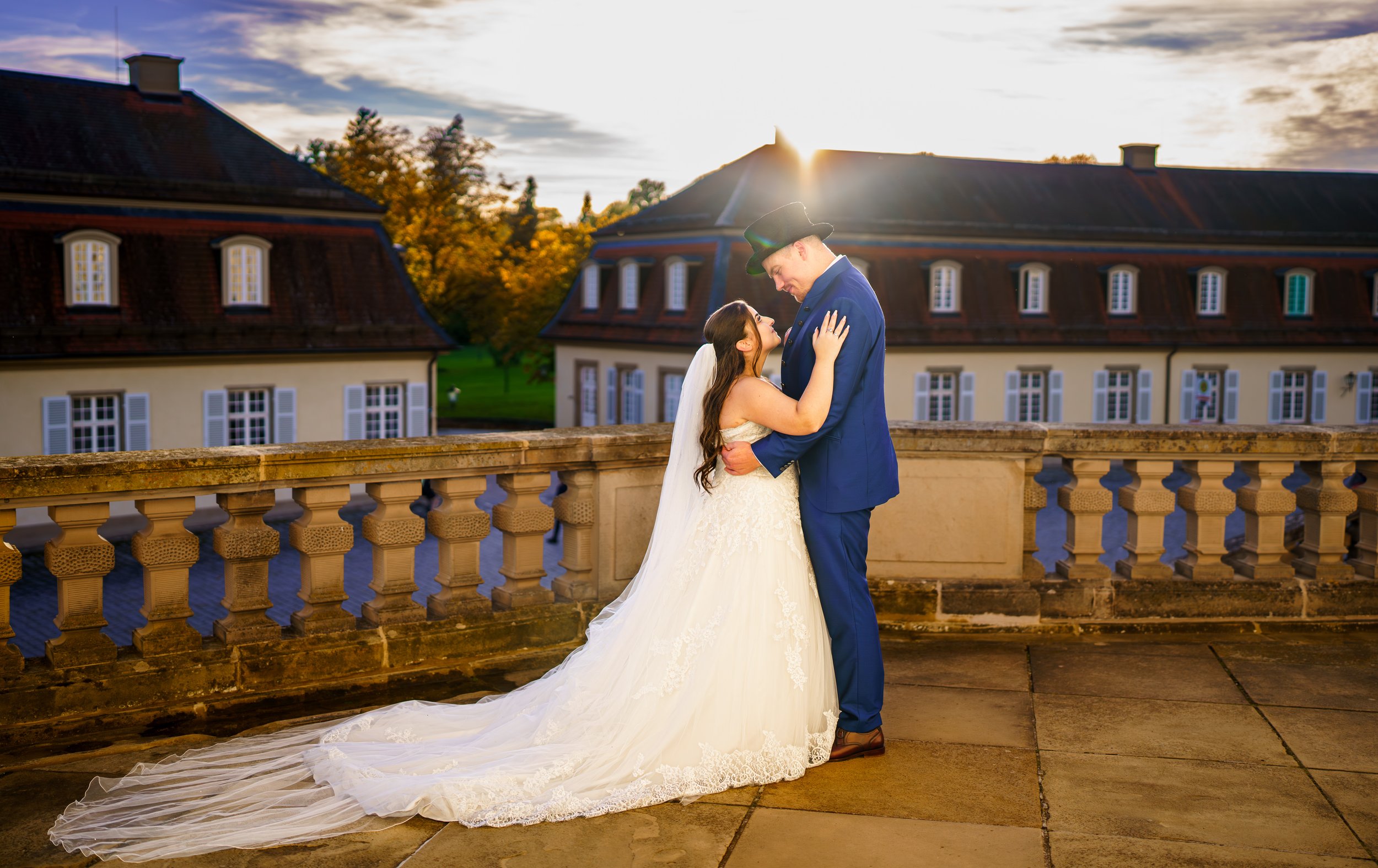 Ein Brautpaar im Hochzeitskleid und Anzug umarmt sich auf einer Terrasse bei Sonnenuntergang, im Hintergrund alte Gebäude und Bäume.