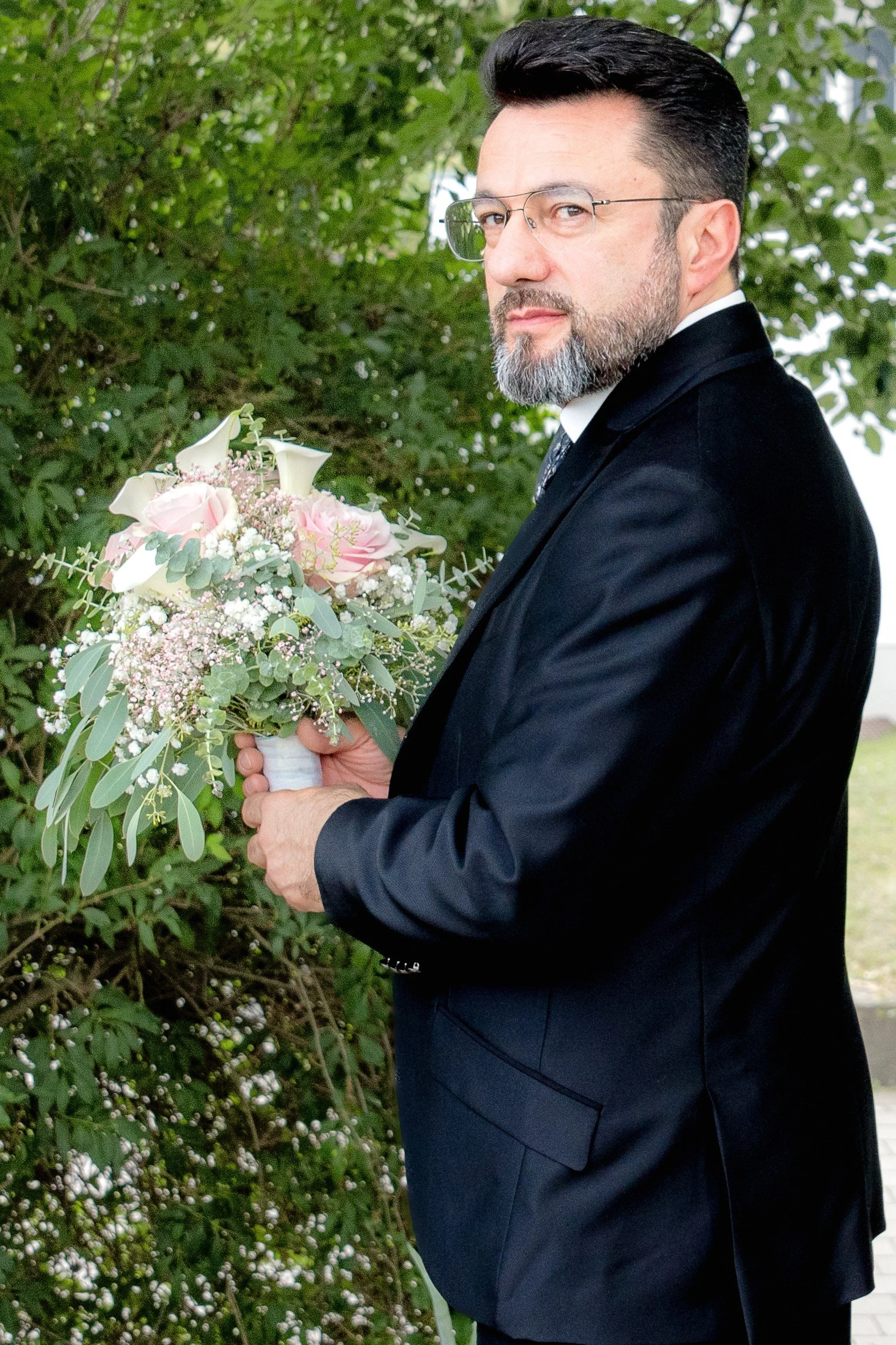 Der Vater kurz vor dem First Look mit seiner Tochter, steht in einem schwarzen Anzug in einem Garten und hält den Brautstrauß  mit rosa Rosen und Calla-Lilien in seiner Hand