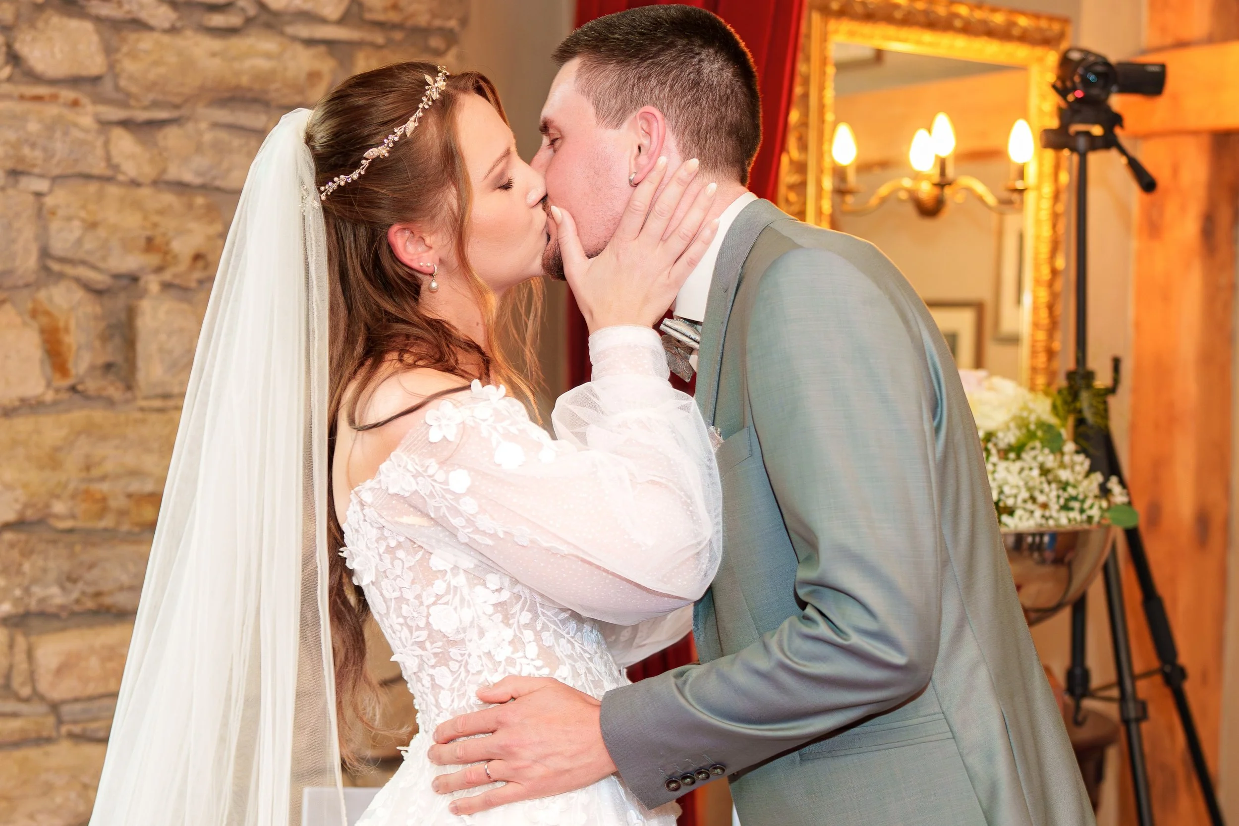 Ein Brautpaar küsst sich während einer Hochzeit, im Hintergrund eine goldene Tapete und ein Gemälde.