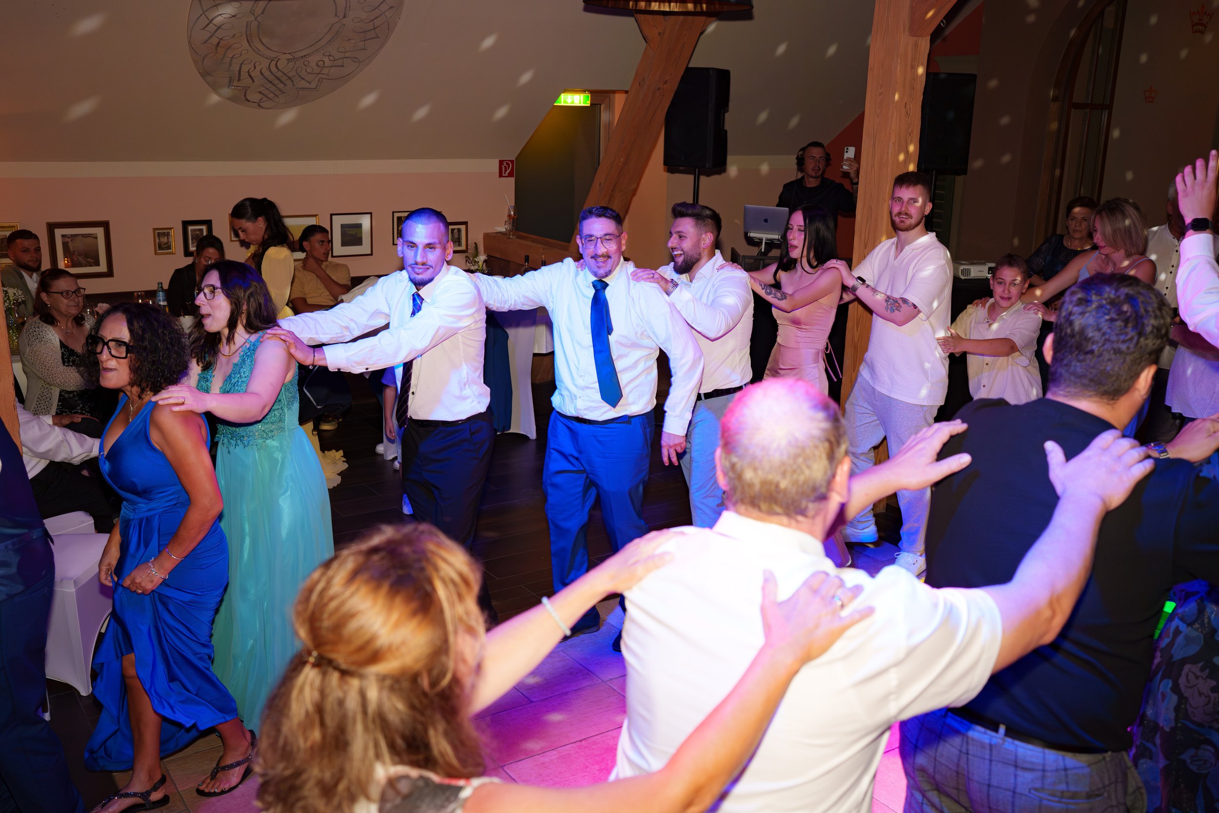 Menschen tanzen in einer Reihe bei einer Feier, alle halten sich an den Schultern und lachen. Die Atmosphäre ist fröhlich, im Hintergrund sitzen Gäste an Tischen, die den Tanz beobachten.