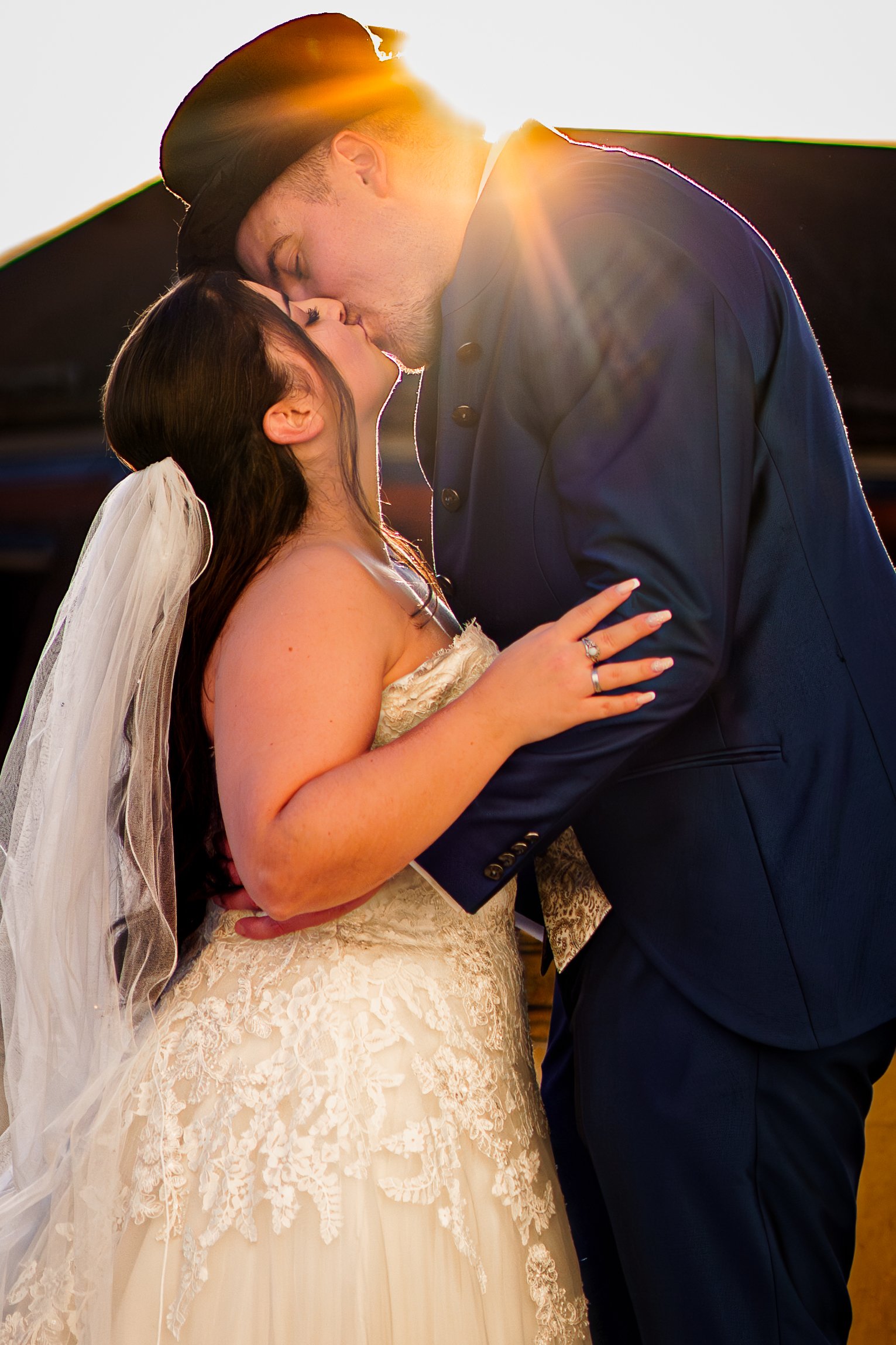 Ein Brautpaar küsst sich bei Sonnenuntergang, die Frau trägt ein weißes Hochzeitskleid mit Spitzendetails und einen Schleier, der Mann trägt einen dunklen Anzug.