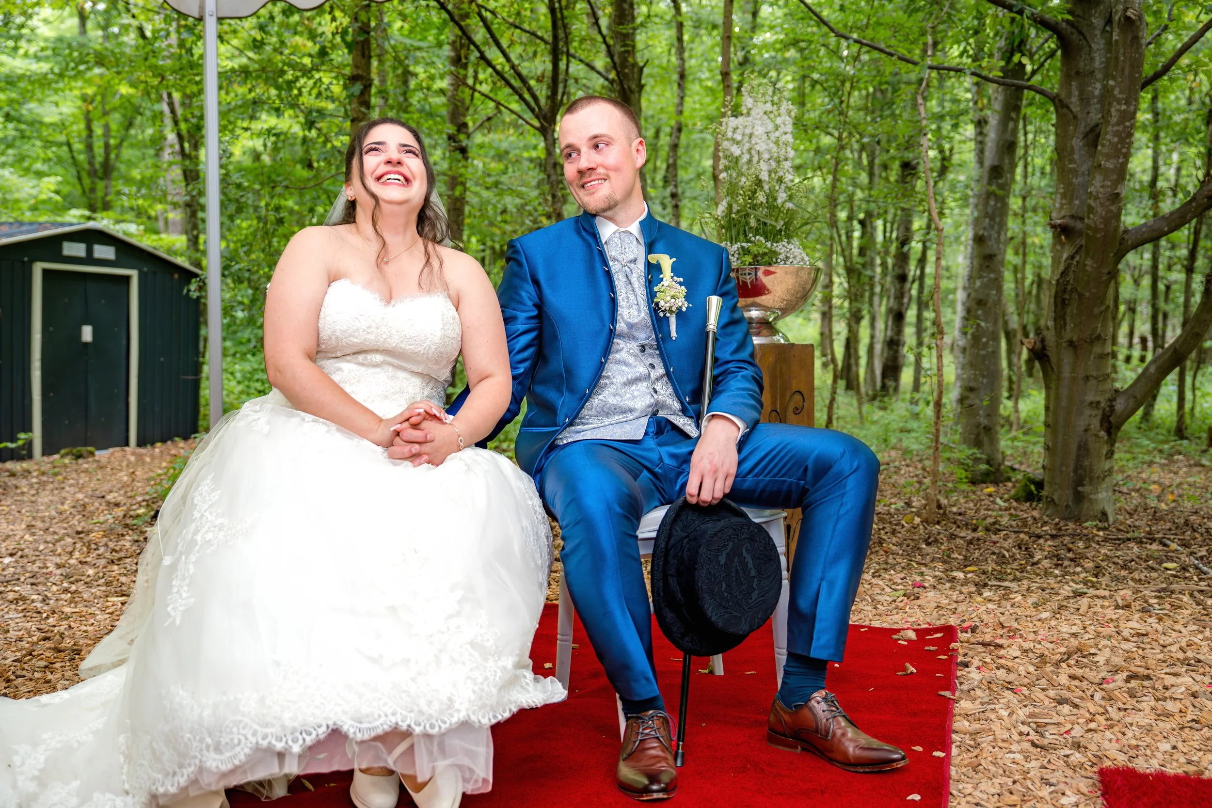 Ein glückliches Brautpaar sitzt auf einem roten Teppich im Wald. Die Braut trägt ein weißes Hochzeitskleid, der Bräutigam einen blauen Anzug und einen Zylinder. Sie halten Händchen und lachen miteinander.