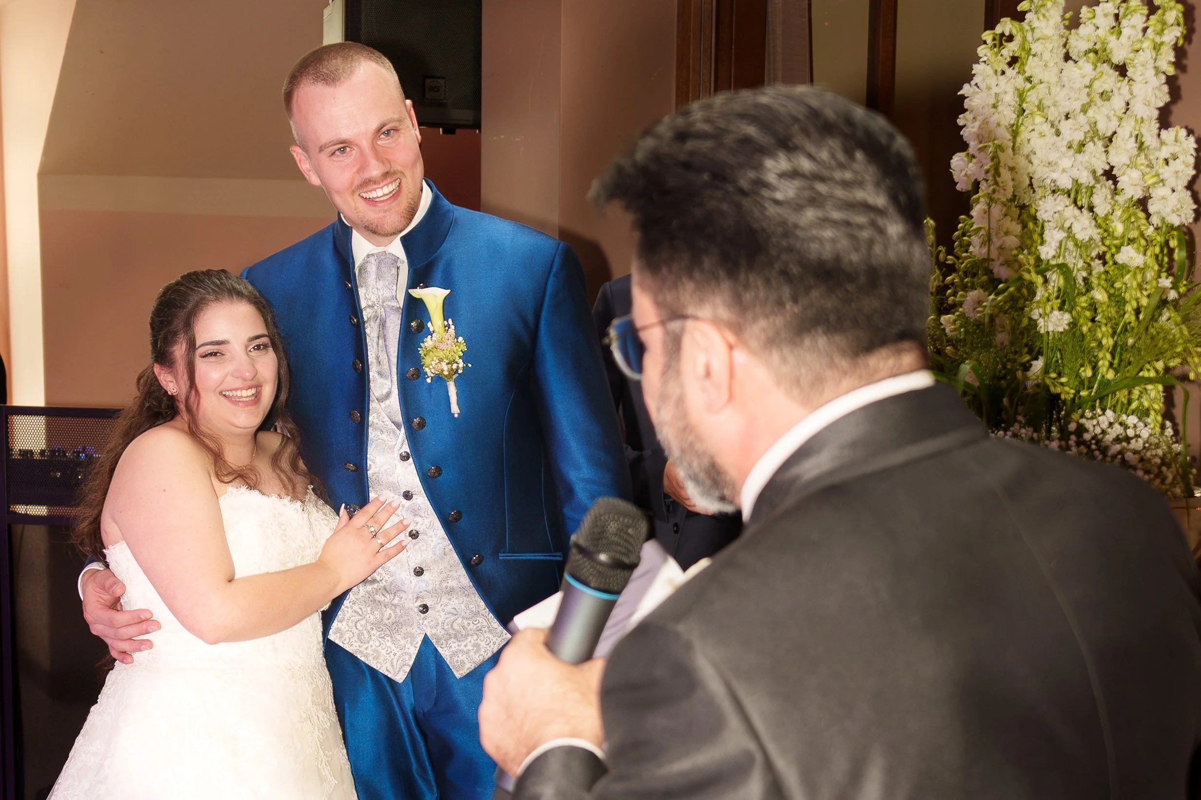 Ein Brautpaar bei ihrer Hochzeit, der Bräutigam in einem blauen Anzug, die Braut in einem weißen Kleid, der Redner mit Mikrofon.