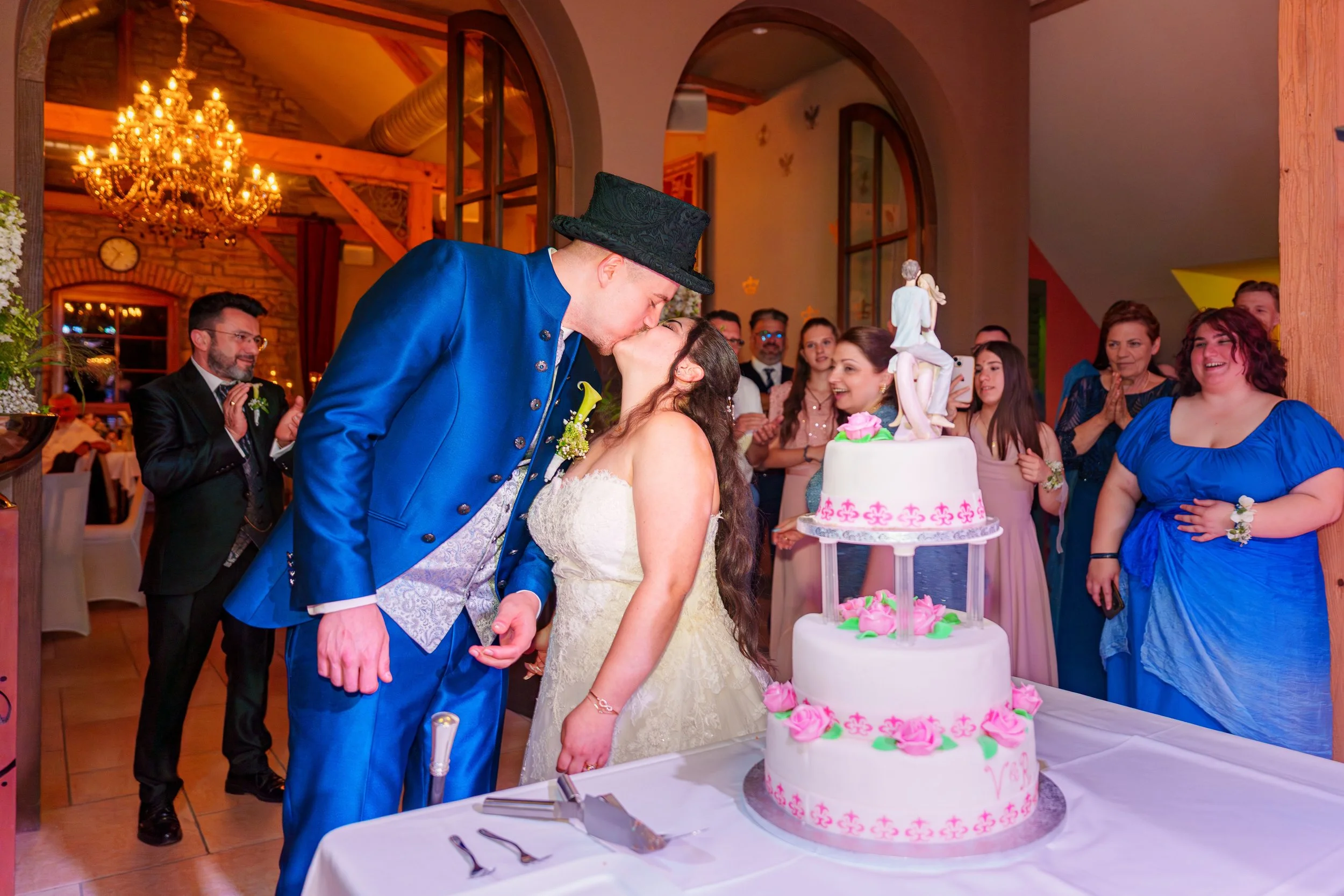 Ein Brautpaar küsst sich vor einer Hochzeitstorte, umgeben von lachenden Gästen in einem festlich dekorierten Raum.