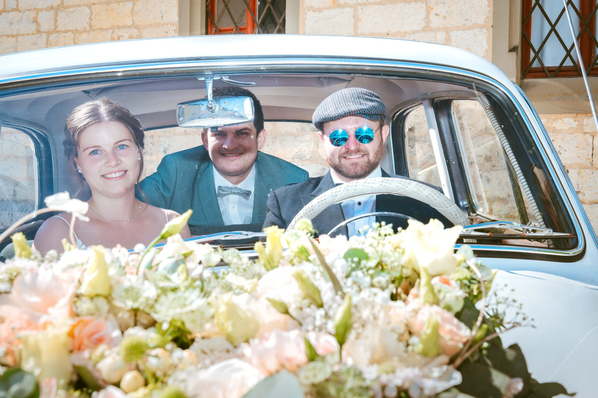 Drei Menschen in einem Oldtimer, eine Frau und zwei Männer, bei einer Hochzeitsfeier, vor einem Blumenarrangement auf der Motorhaube.