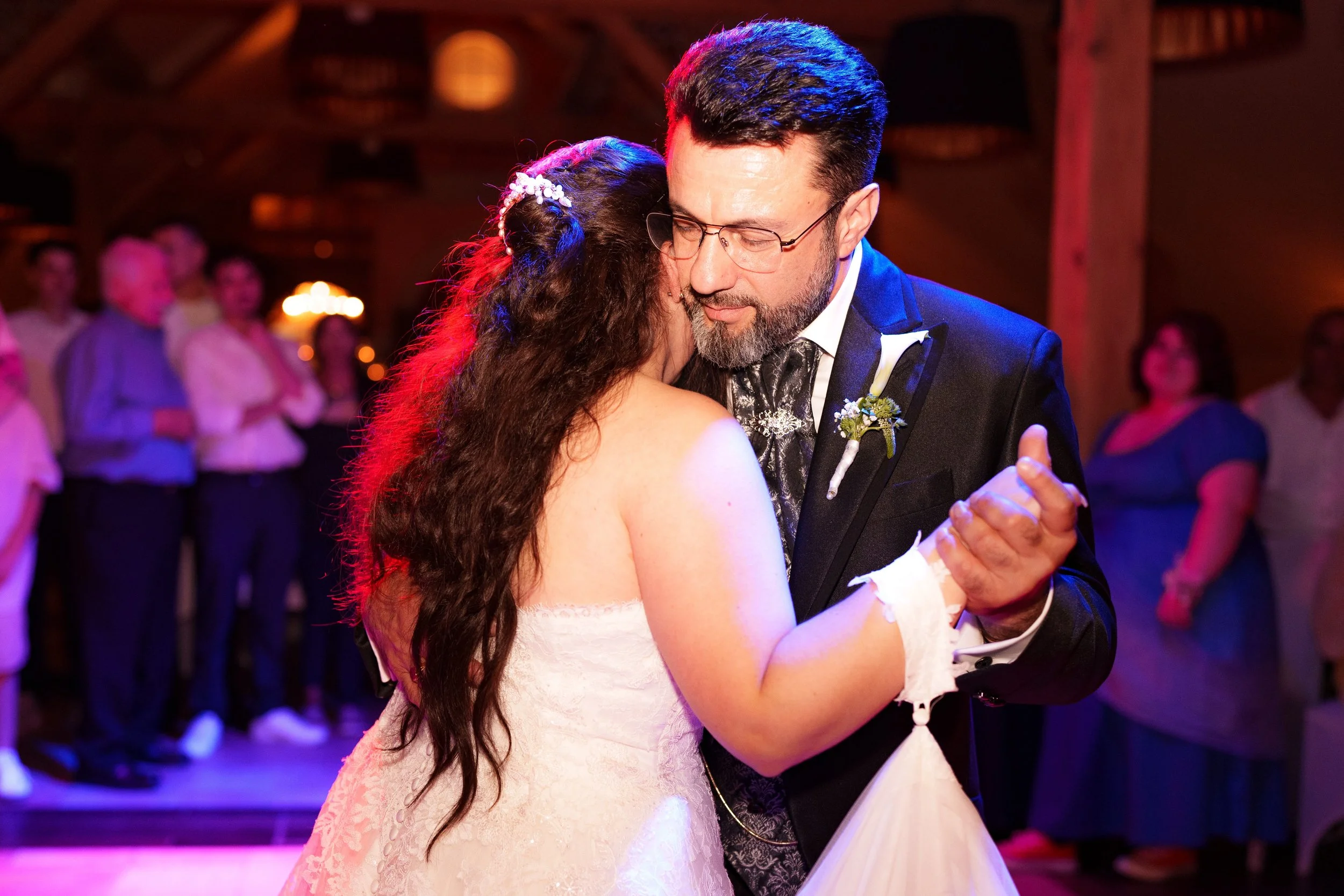 Ein Brautpaar tanzt bei einer Hochzeit, im Hintergrund stehen Gäste, die die Feier beobachten.