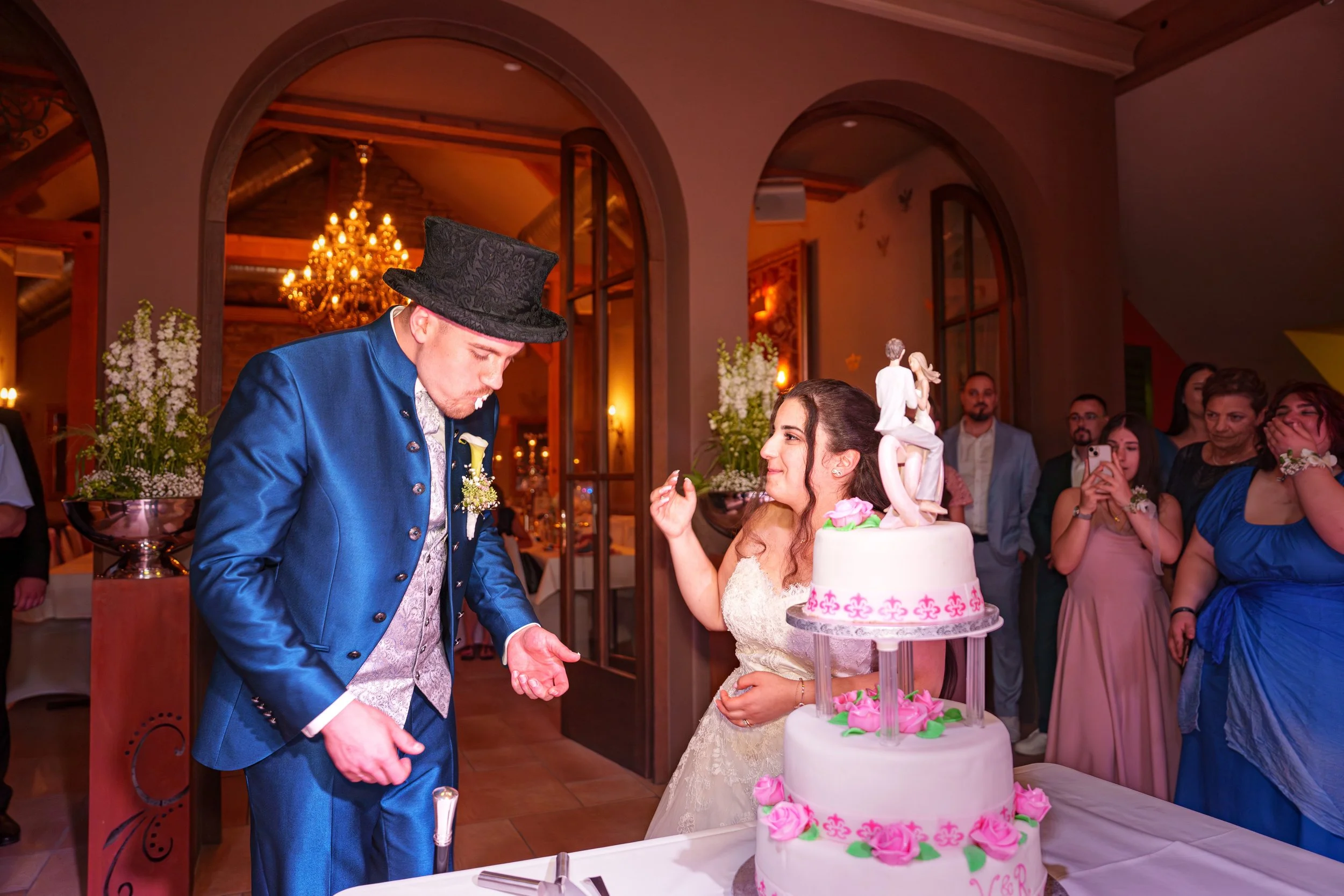 Ein Hochzeitspaar mit Kuchen und Gästen im Hintergrund in einem festlich dekorierten Raum.