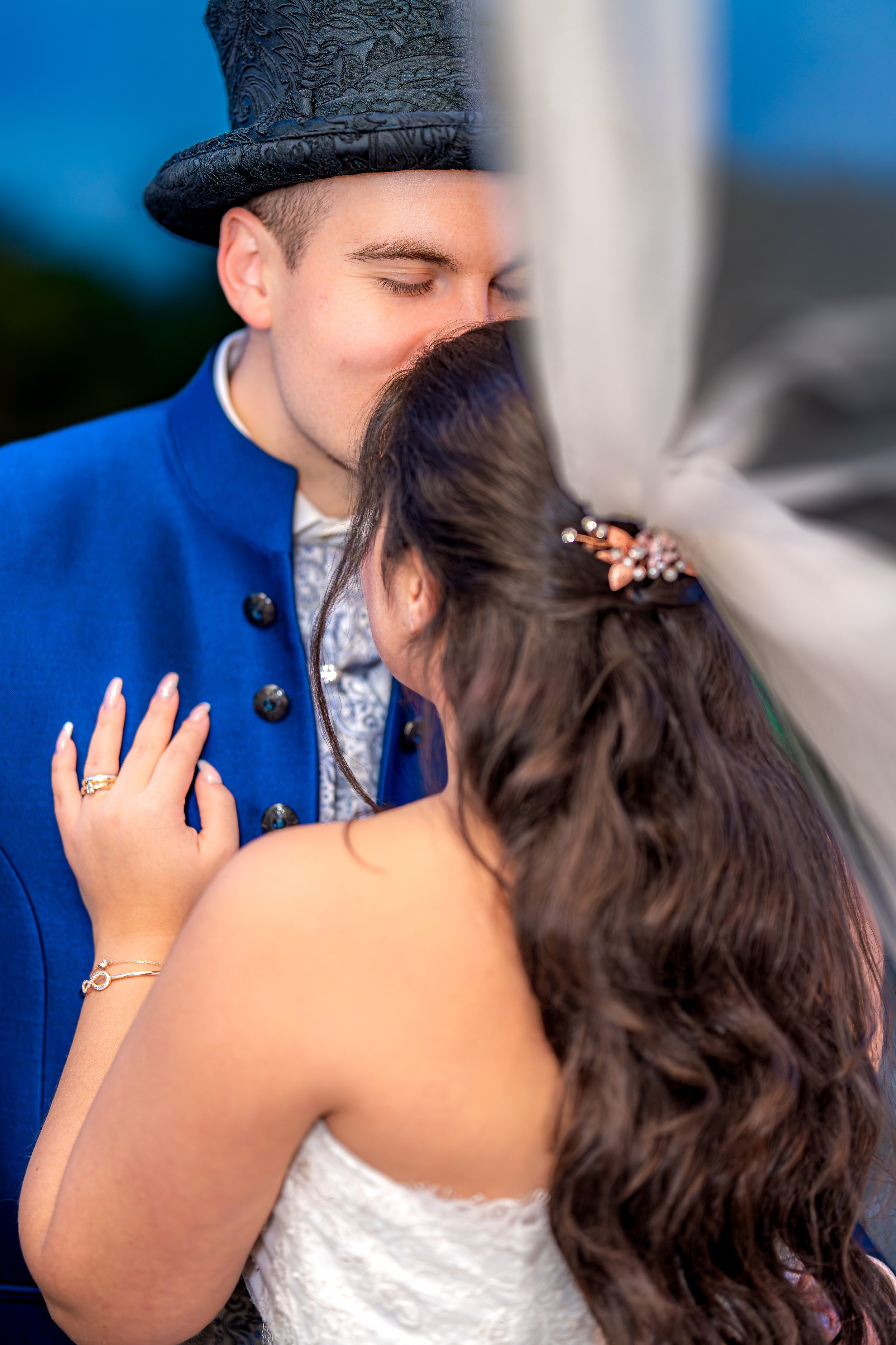 Braut und Bräutigam küssen sich an ihrer Hochzeit, der Bräutigam trägt einen blauen Anzug und einen schwarzen Zylinder, die Braut trägt ein weißes Kleid mit langen, lockigen Haaren und Haaraccessoire.