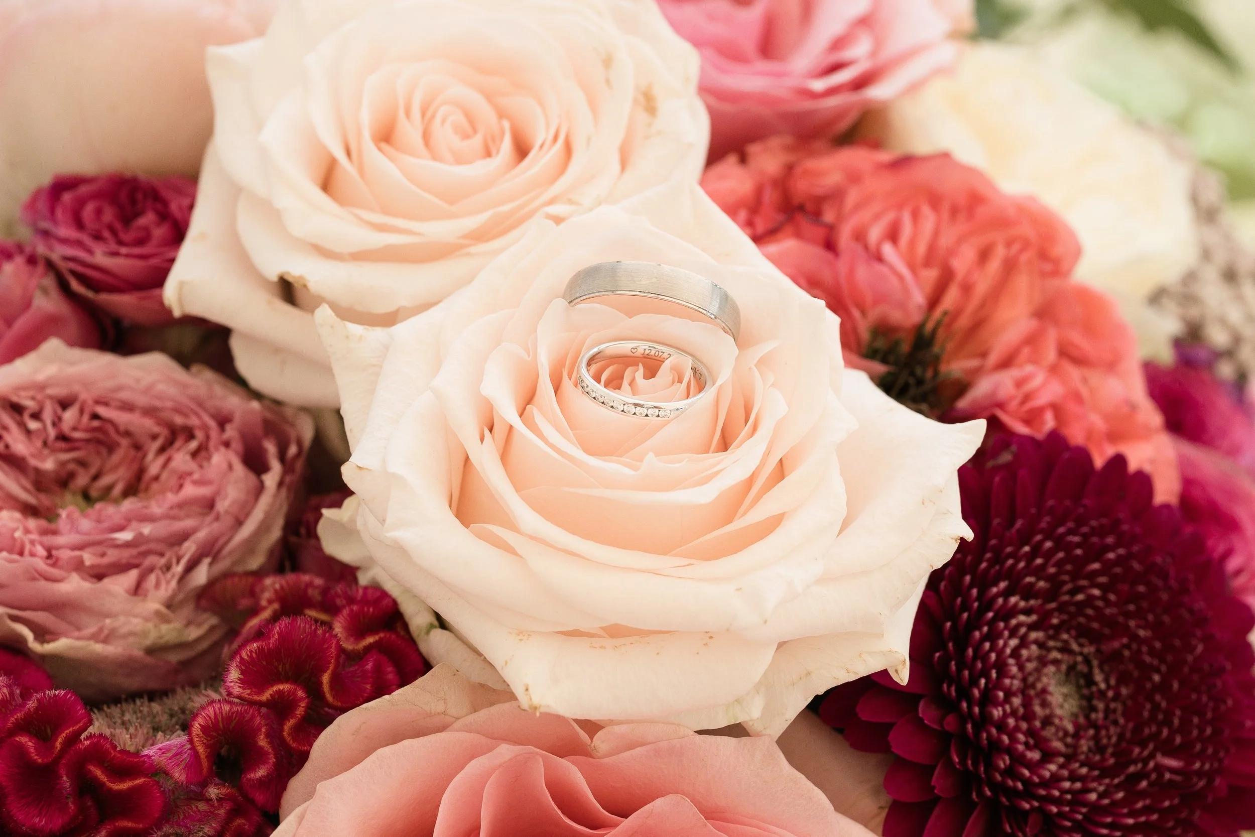 Verlobungs- und Eheringe liegen auf einer weißen Rose inmitten eines Blumenbouquets aus verschiedenen rosafarbenen und roten Blumen.