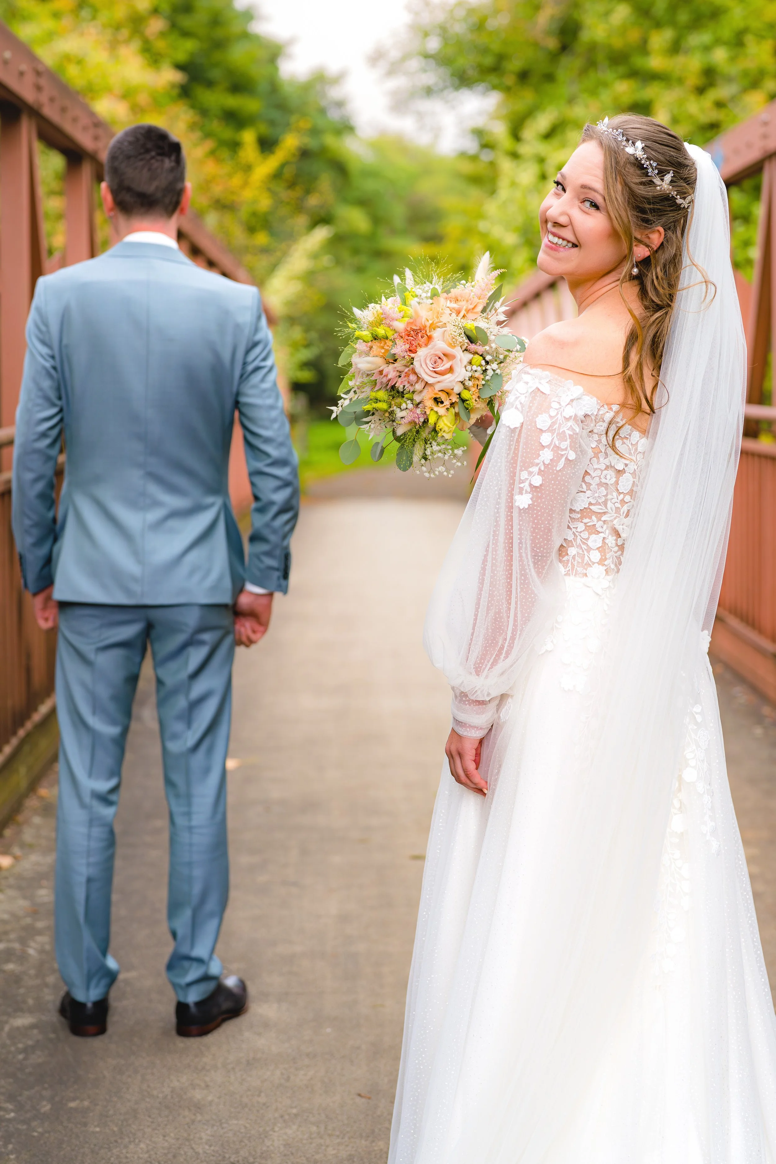 Braut, die in einem weißen Hochzeitskleid und mit Blumenstrauß lächelnd auf dem Weg zum Bräutigam steht, während der Bräutigam mit Rückenansicht auf einer Brücke steht.