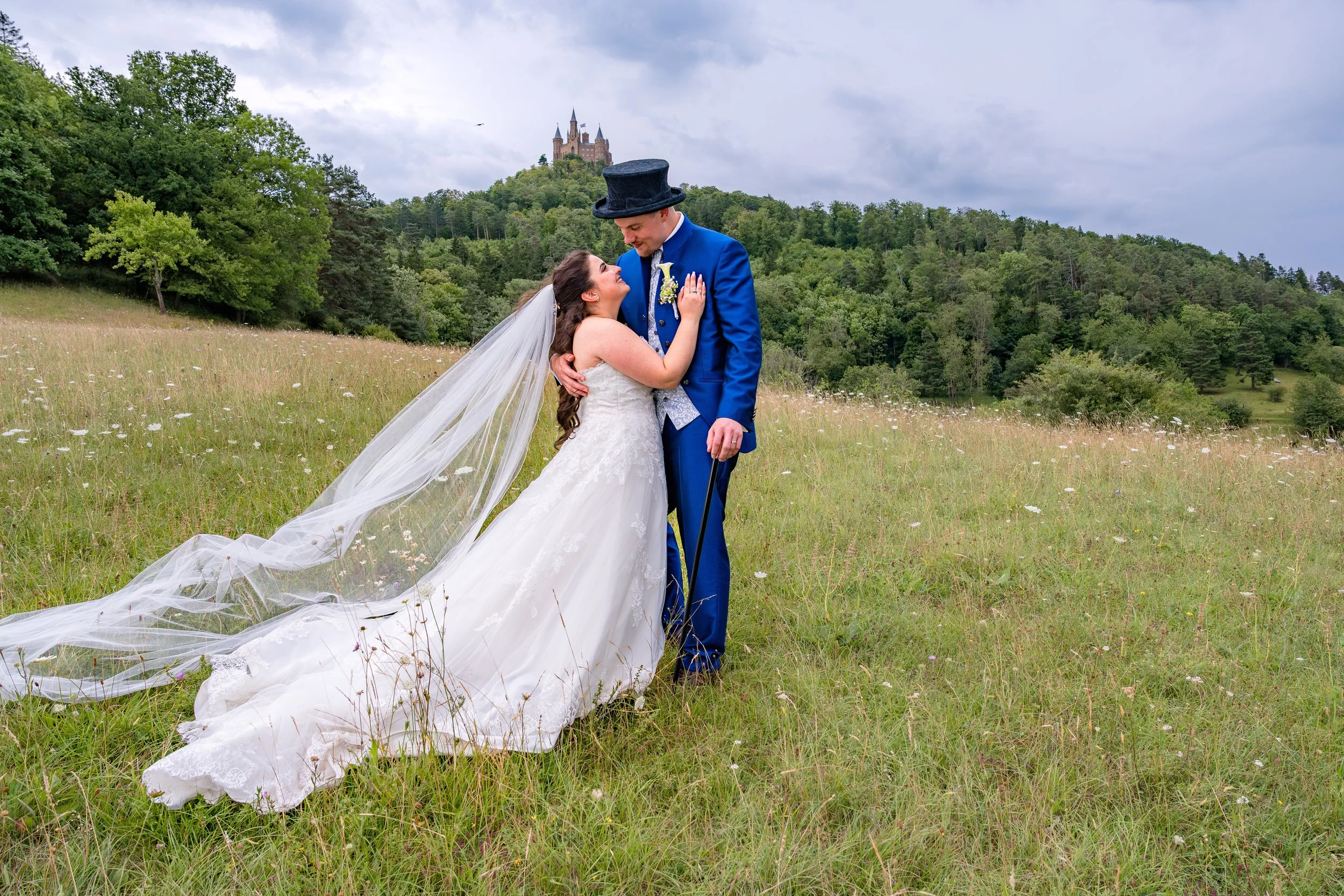 Braut und Bräutigam im Hochzeitsoutfit auf einer Wiese mit Schloss im Hintergrund, umarmend und lächelnd.