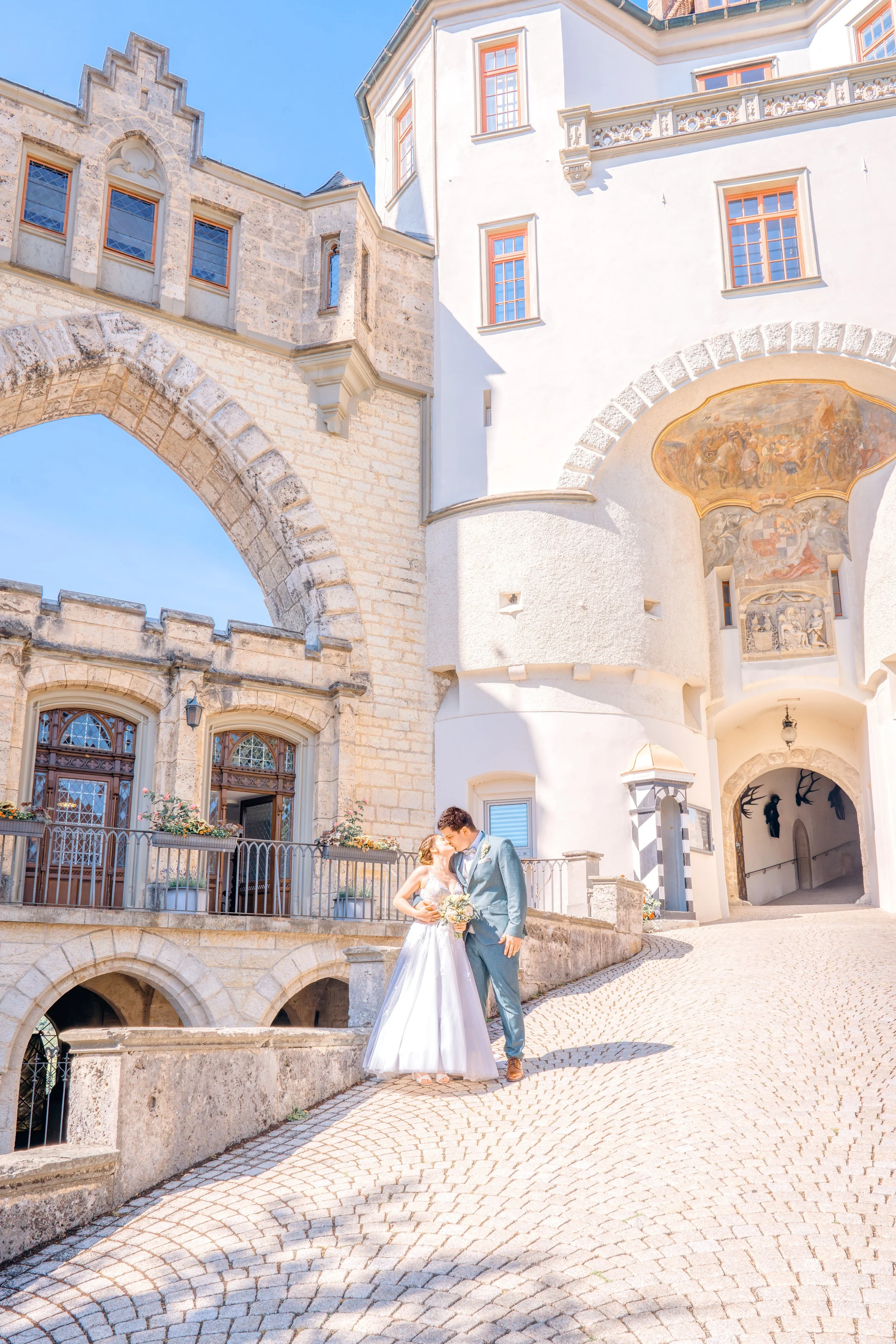 Ein Brautpaar steht vor einem historischen Gebäude mit hellem Putz und Steingebäudeteilen. Das Paar umarmt sich liebevoll, die Braut trägt ein weißes Hochzeitskleid, der Bräutigam einen hellblauen Anzug. Das Gebäude hat bunte Fenster und kunstvolle W