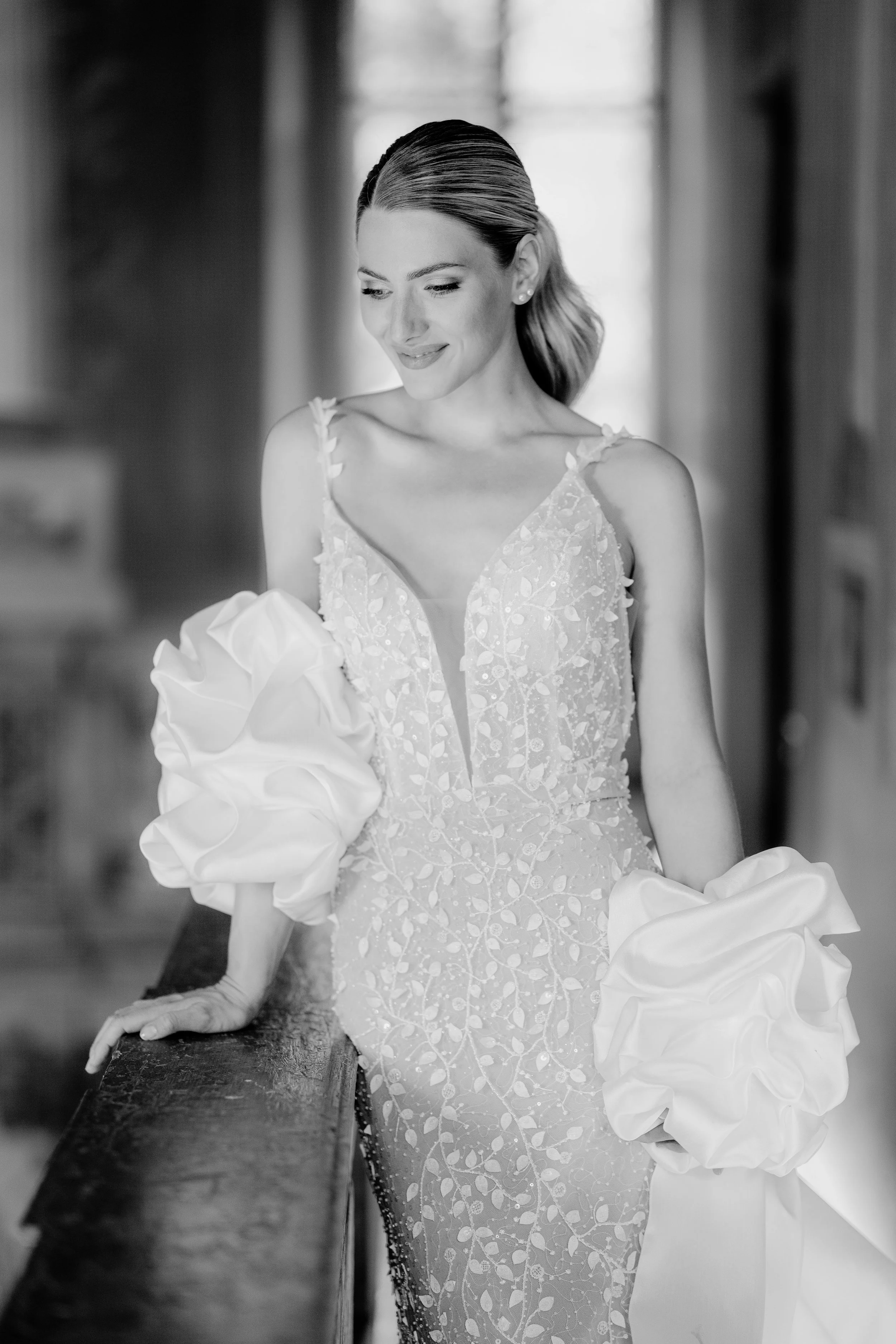 Eine Frau in einem eleganten Hochzeitskleid, die an einem Handlauf lehnt und nach unten schaut, in einem Gang mit natürlichem Licht.