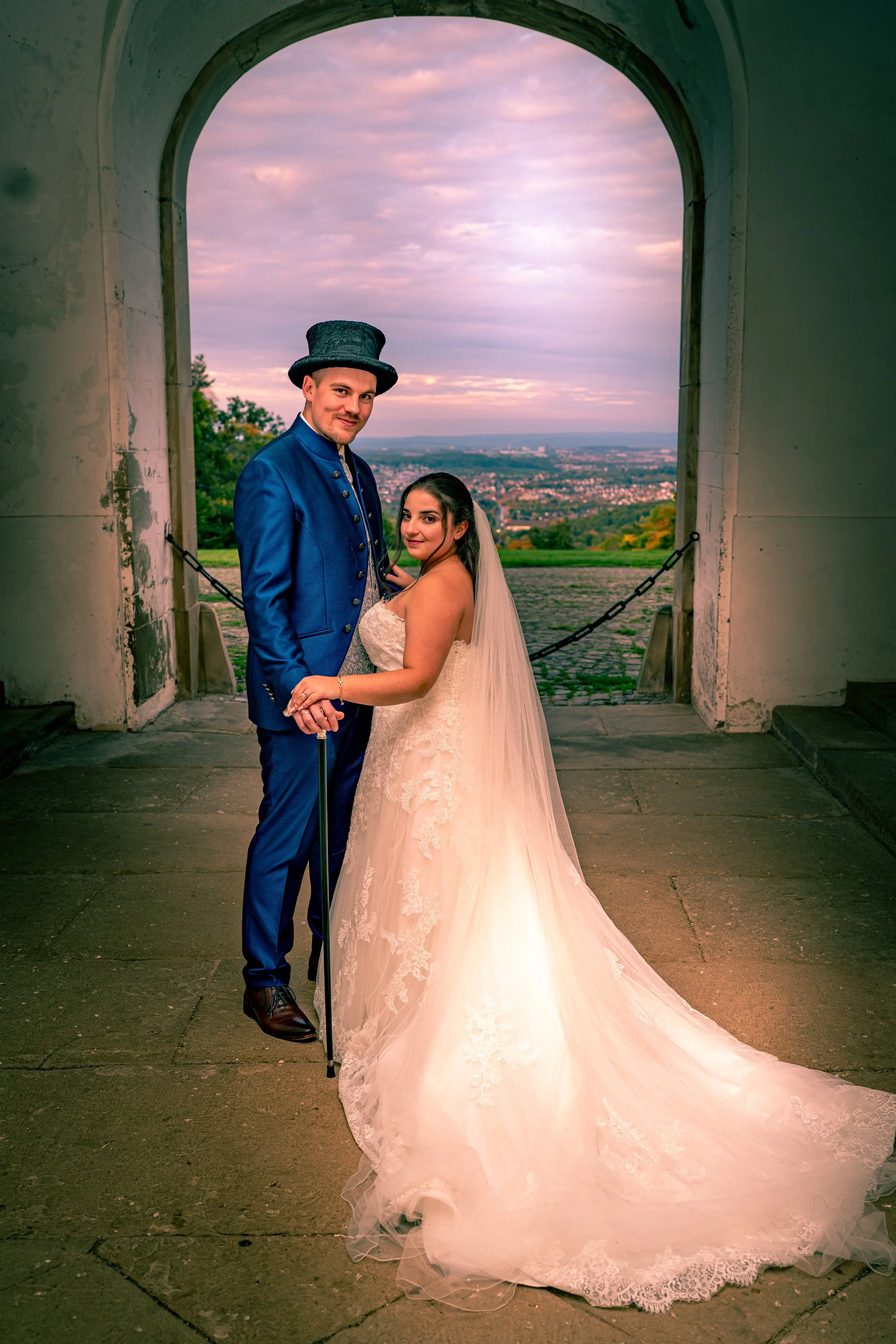 Ein Brautpaar in Hochzeitskleidung steht unter einem großen, alten Torbogen mit Blick auf eine Stadt und die Landschaft bei Sonnenuntergang.