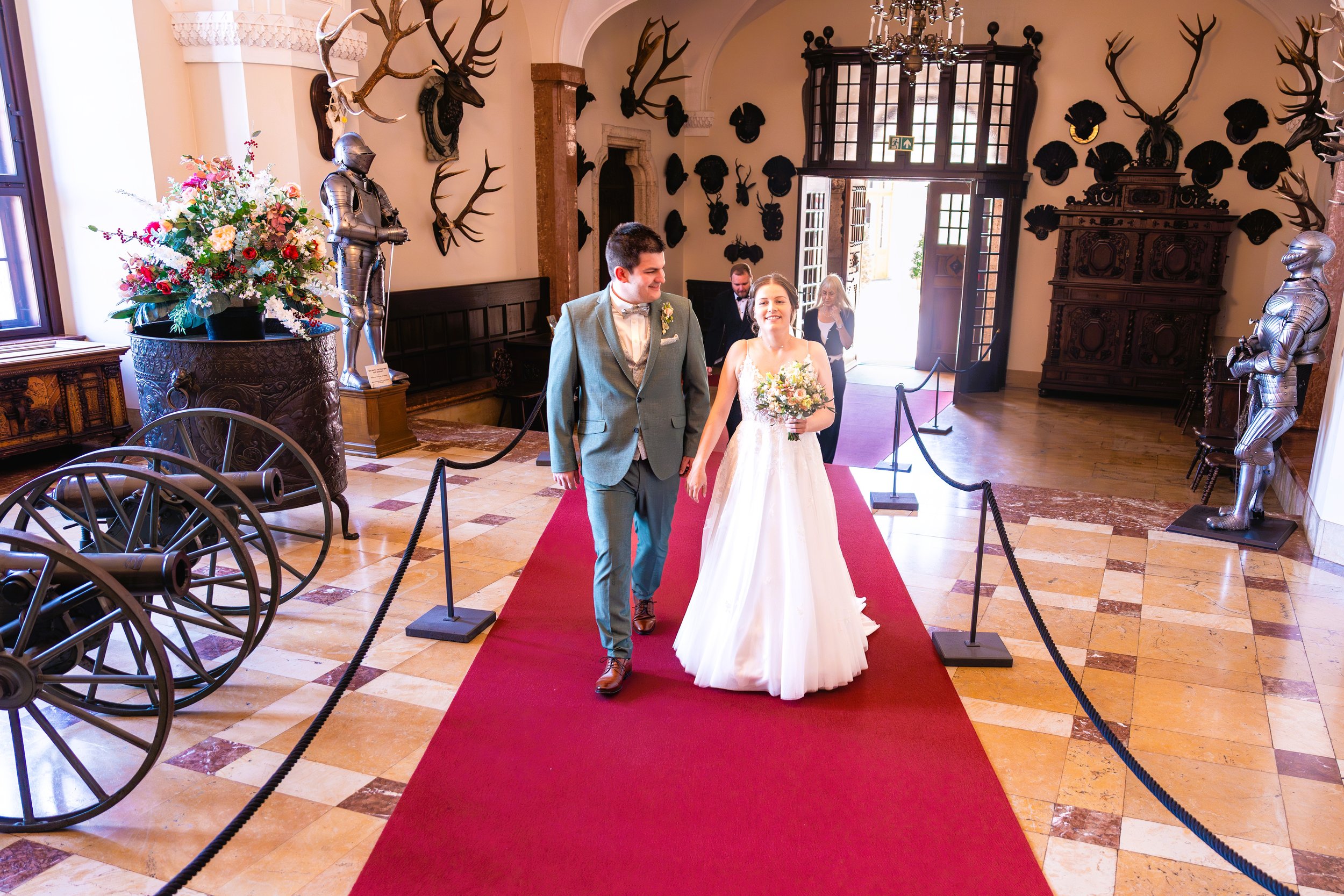 Ein frisch vermähltes Brautpaar läuft die Rotte am Eingang einer historischen, reich verzierten Schloss-Halle entlang, umgeben von Deko mit Ritterrüstungen und Jagdtrophäen.