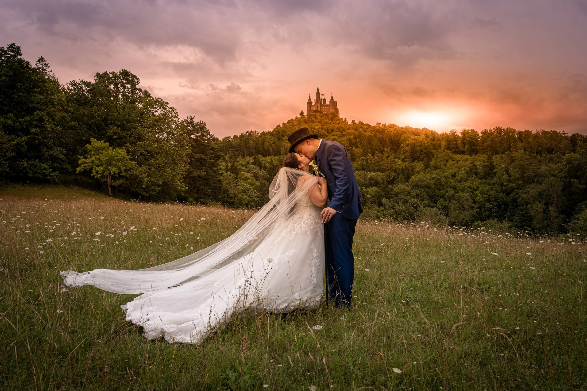 Ein Brautpaar küsst sich in einem Feld mit einer Burg im Hintergrund bei Sonnenuntergang.