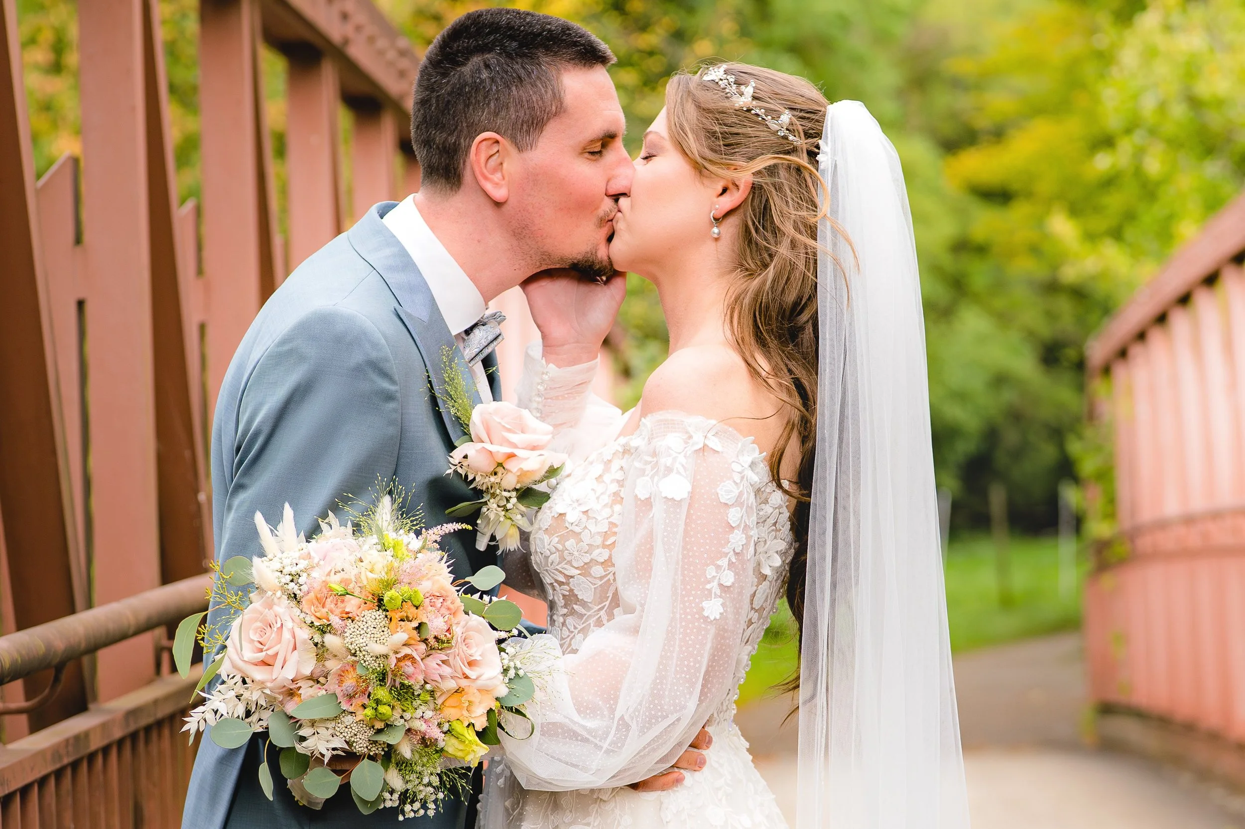 Ein Brautpaar küsst sich vor einem orangefarbenen Zaun in einer grünen Parklandschaft.