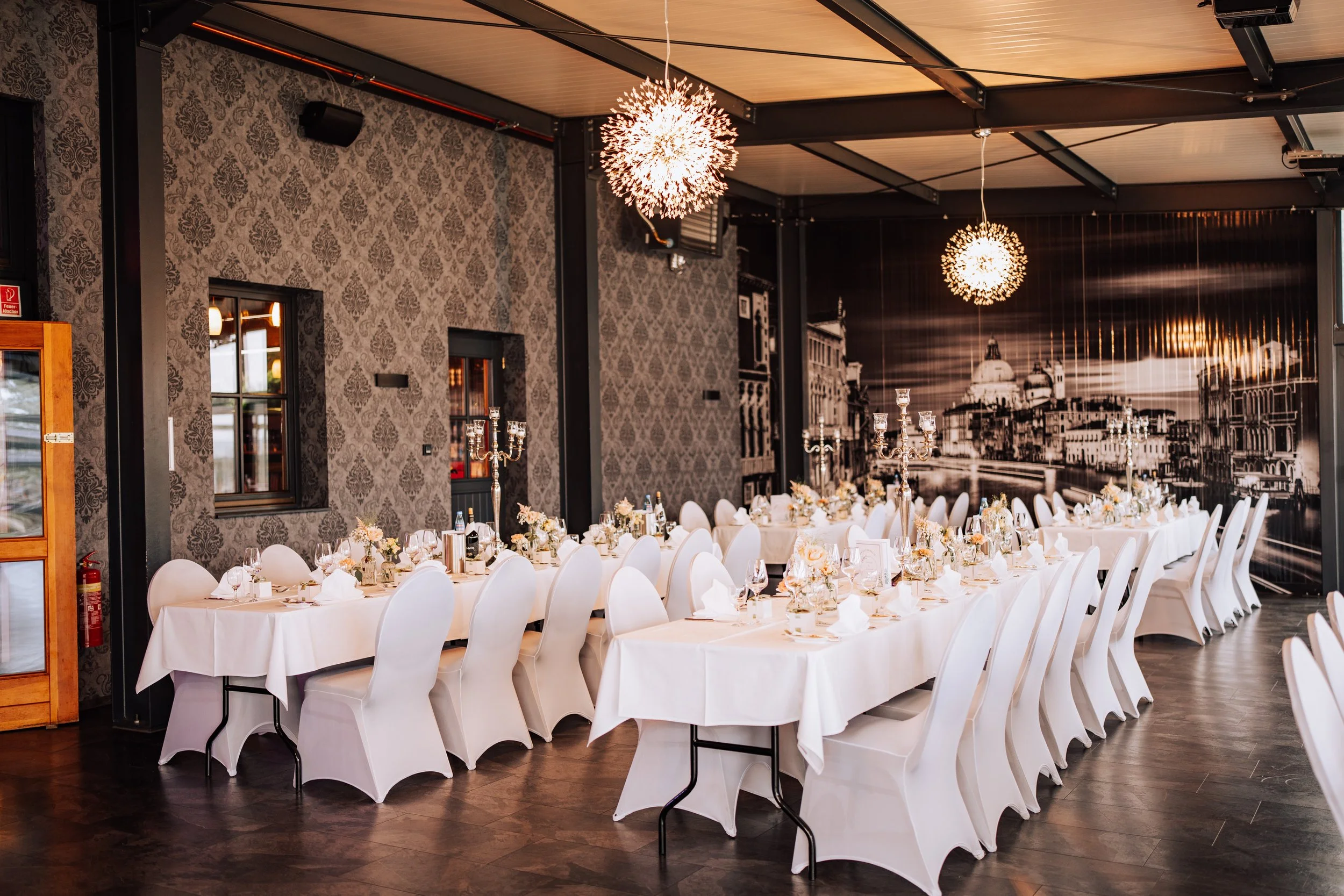 Ein festlich gedeckter Veranstaltungsraum mit langen Tischen, weißer Tischwäsche, Kerzen, Glaswaren und goldenen Blumenarrangements, mit moderner Beleuchtung und Wanddekoration im Hintergrund.